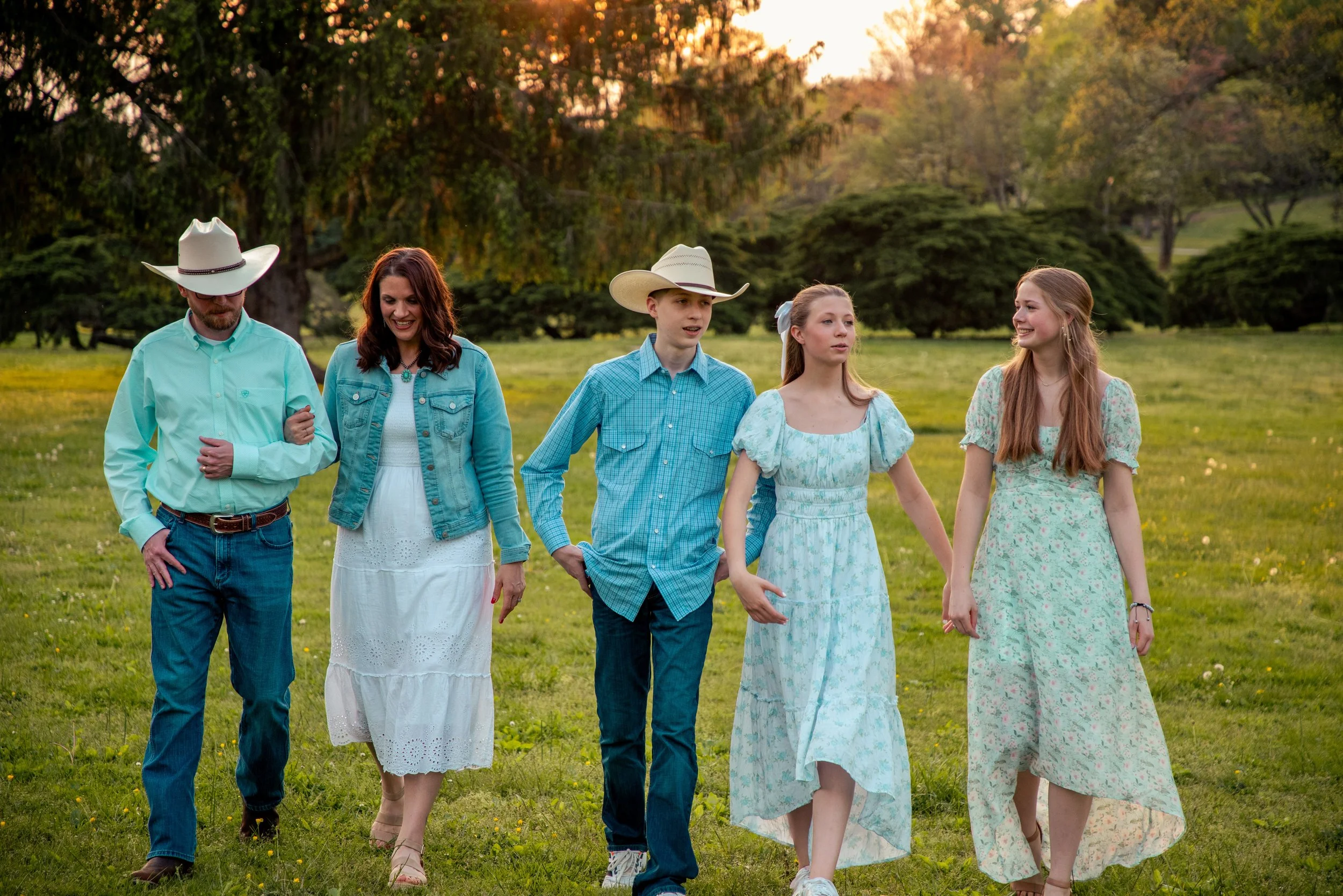 A group of five people, three girls and two boys, walking through a grassy park during sunset. The adults are wearing cowboy hats, and the girls are dressed in pastel-colored dresses.