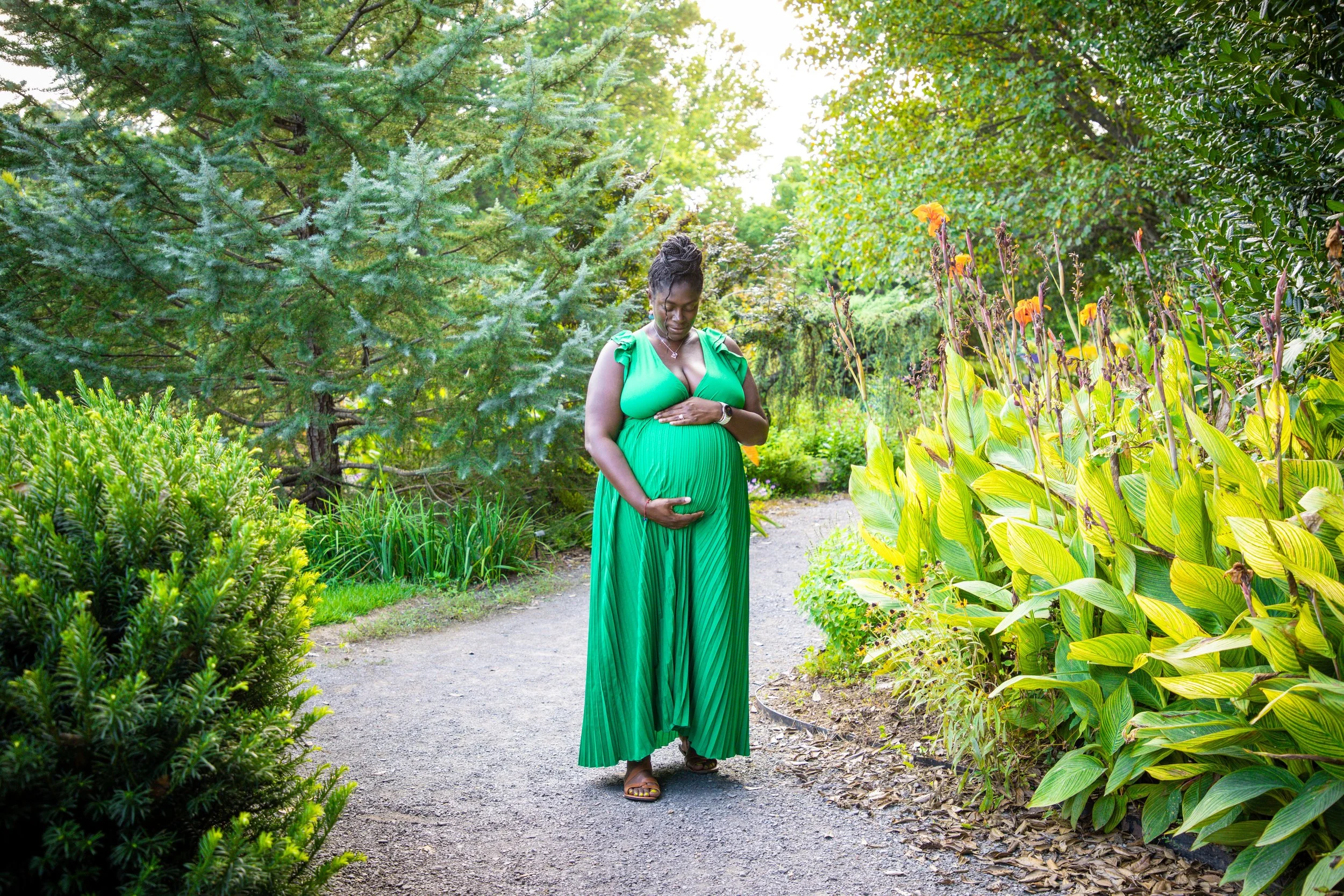Pregnant woman in a green dress holding her belly while standing on a nature trail surrounded by green plants and trees.