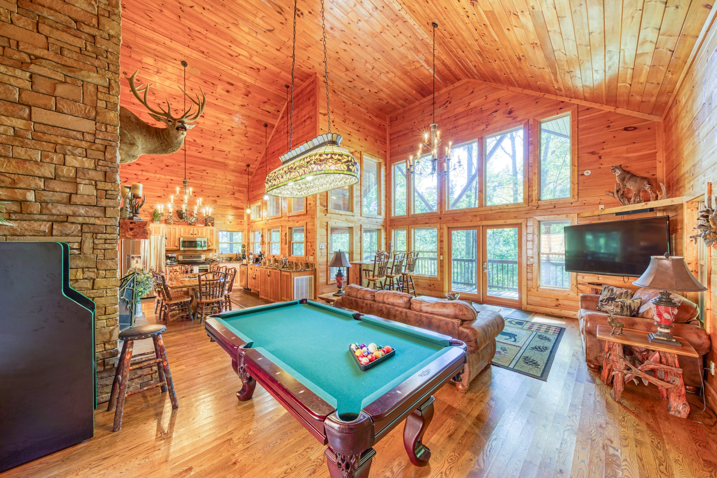 Interior of a wooden cabin living room with large windows, a stone fireplace with mounted deer head, a pool table, a couch, a television, and taxidermy animals on the shelves.
