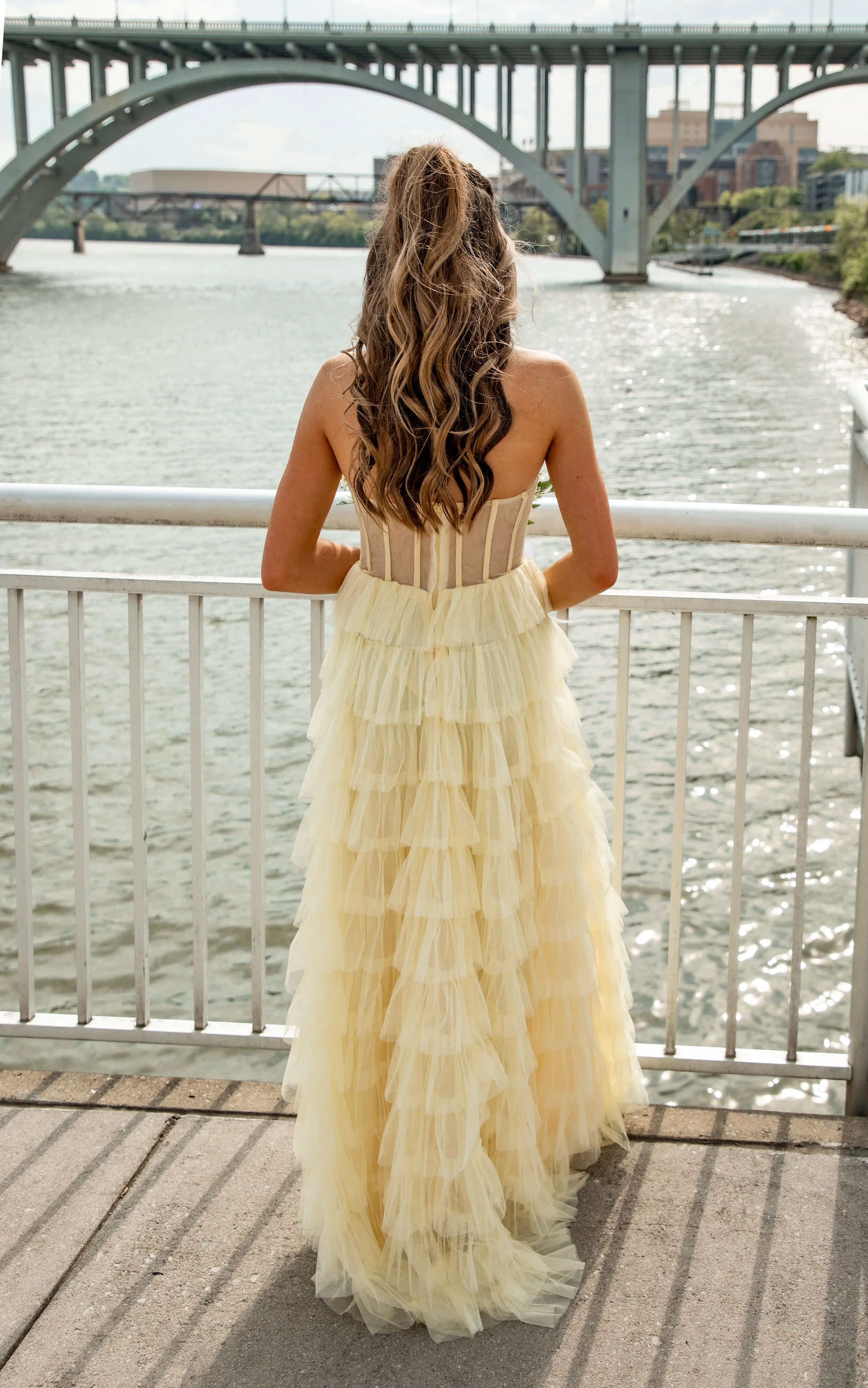 Woman in a yellow dress with layered tulle skirt stands on a riverside walkway, facing away, with bridges and city buildings in the background.