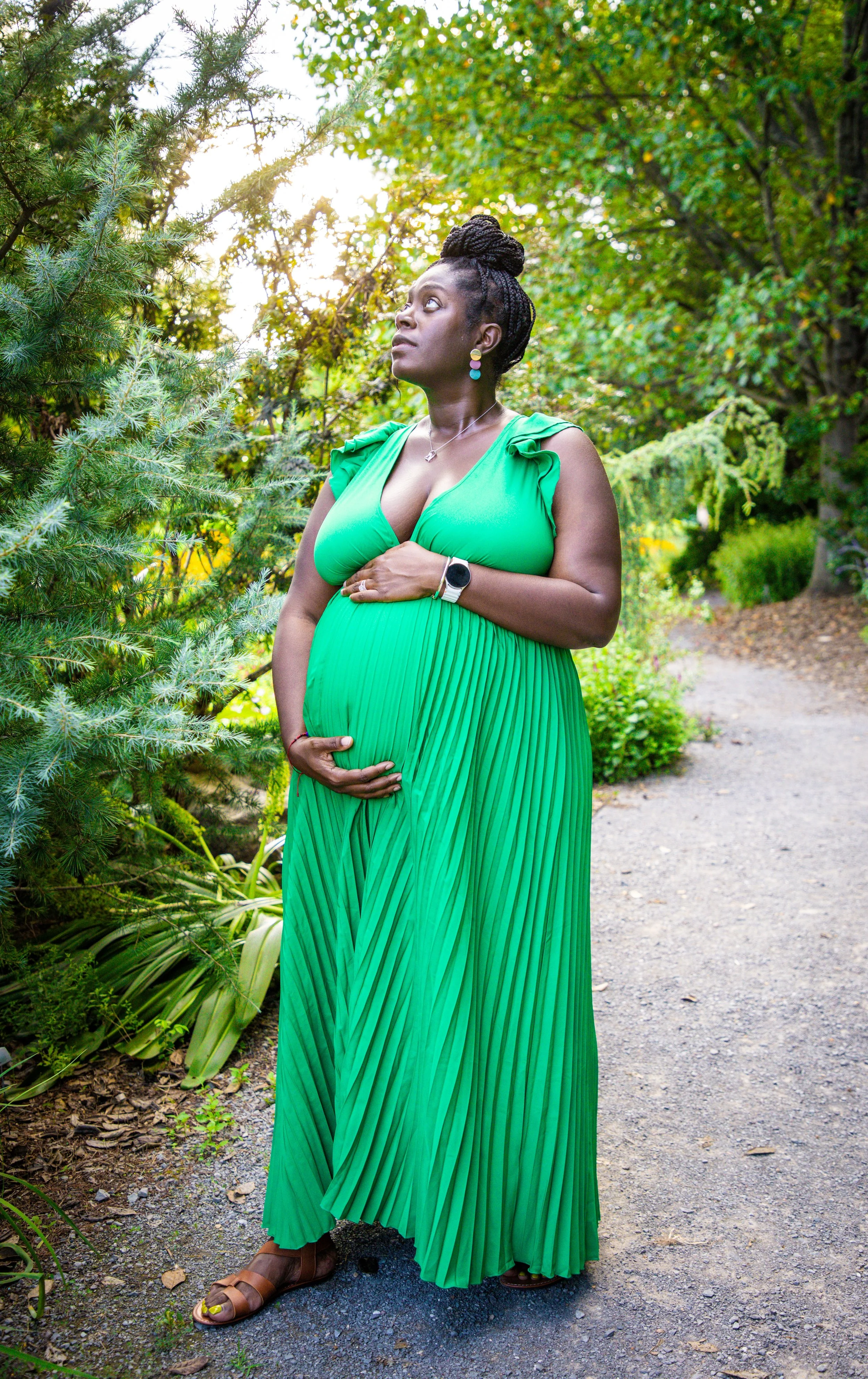 A woman in a green pleated maxi dress with ruffle details on the shoulders, standing in a lush, green garden with trees and bushes, holding her stomach, looking up and to the left.