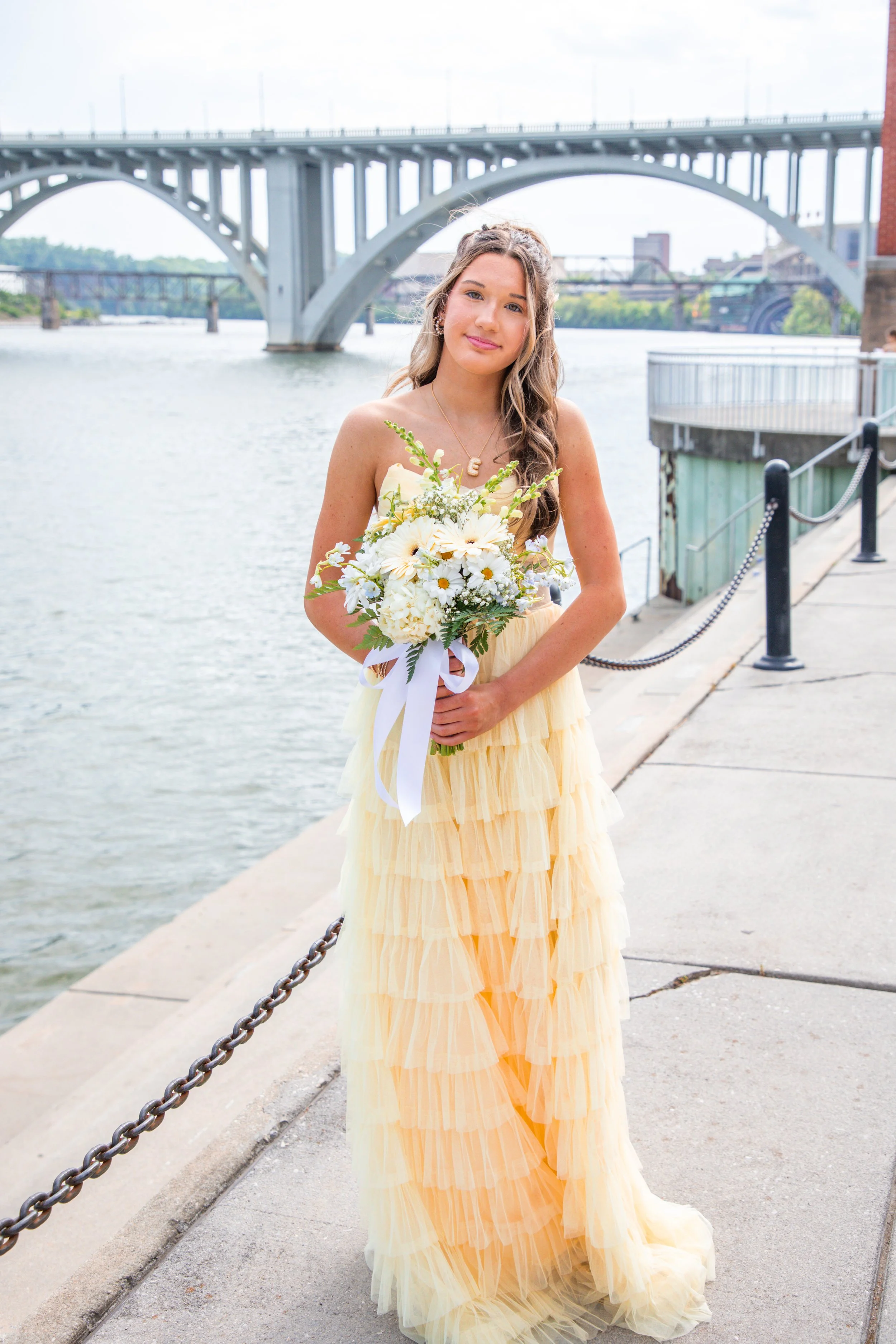 Young woman in yellow dress holding a bouquet of white flowers near a waterfront with a bridge in the background.