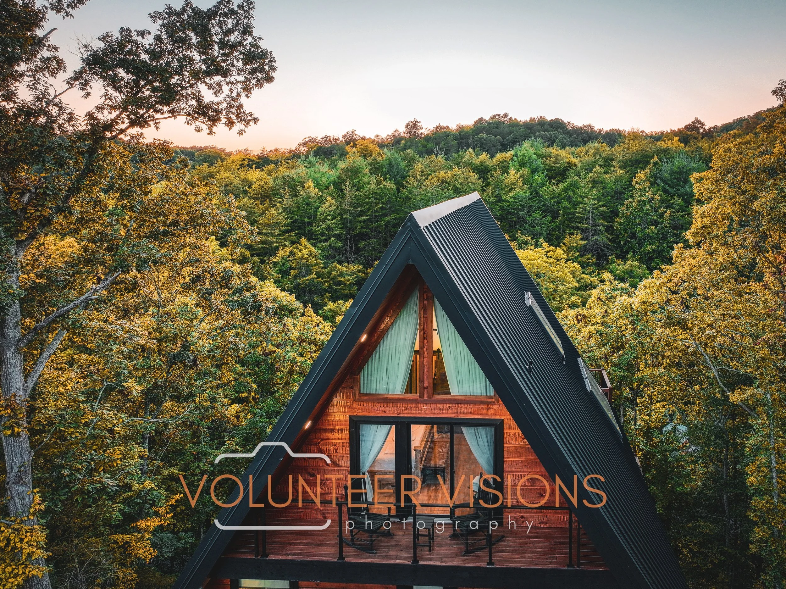 A glass-fronted A-frame building with a black metal roof and wooden walls, situated among a dense forest of green and yellow autumn trees, during sunset.