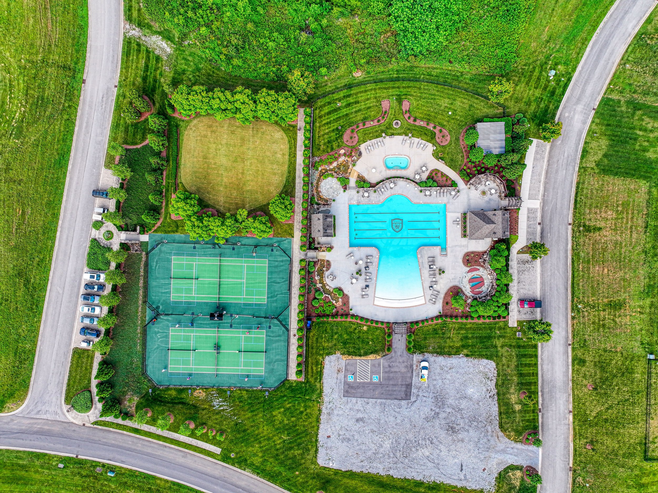 An aerial view of a residential recreational area featuring a tennis court, a swimming pool, a playground, a grassy field, and surrounding roads and trees.