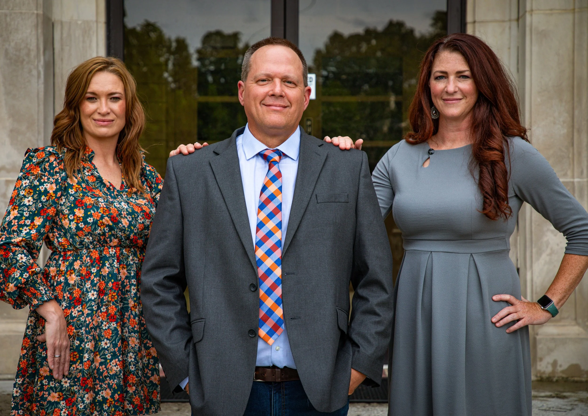Three adults standing outdoors in front of a building with glass doors. The woman on the left has shoulder-length wavy red hair and is wearing a colorful floral dress. The man in the middle has short light brown hair and is dressed in a gray suit wit