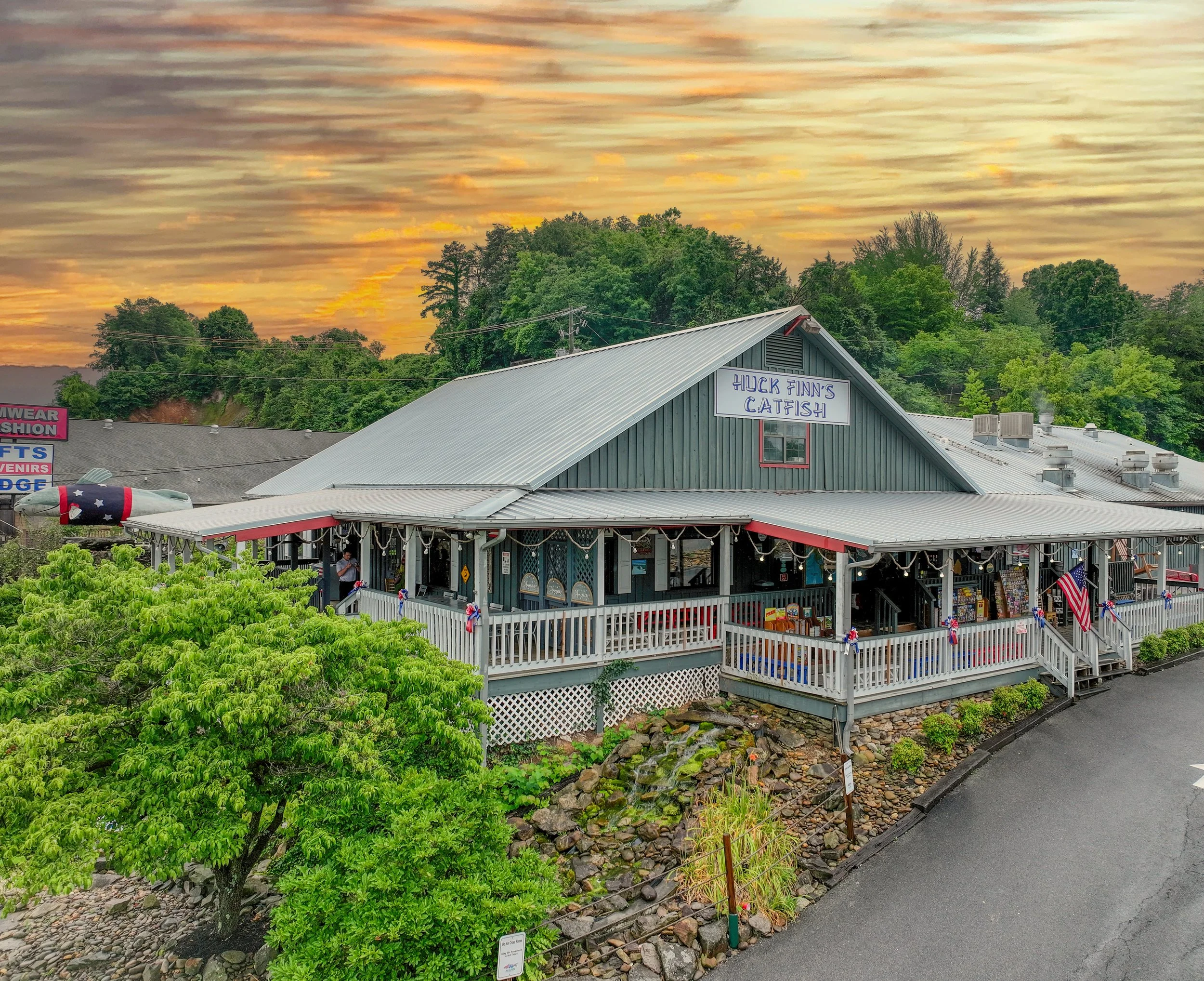 A scenic view of a grey building with a sign that reads 'Huck Finn's Catfish' under a colorful sunset sky. The building has a white porch with tables, a fence decorated with patriotic bunting, and is surrounded by lush green trees and a rocky landsca