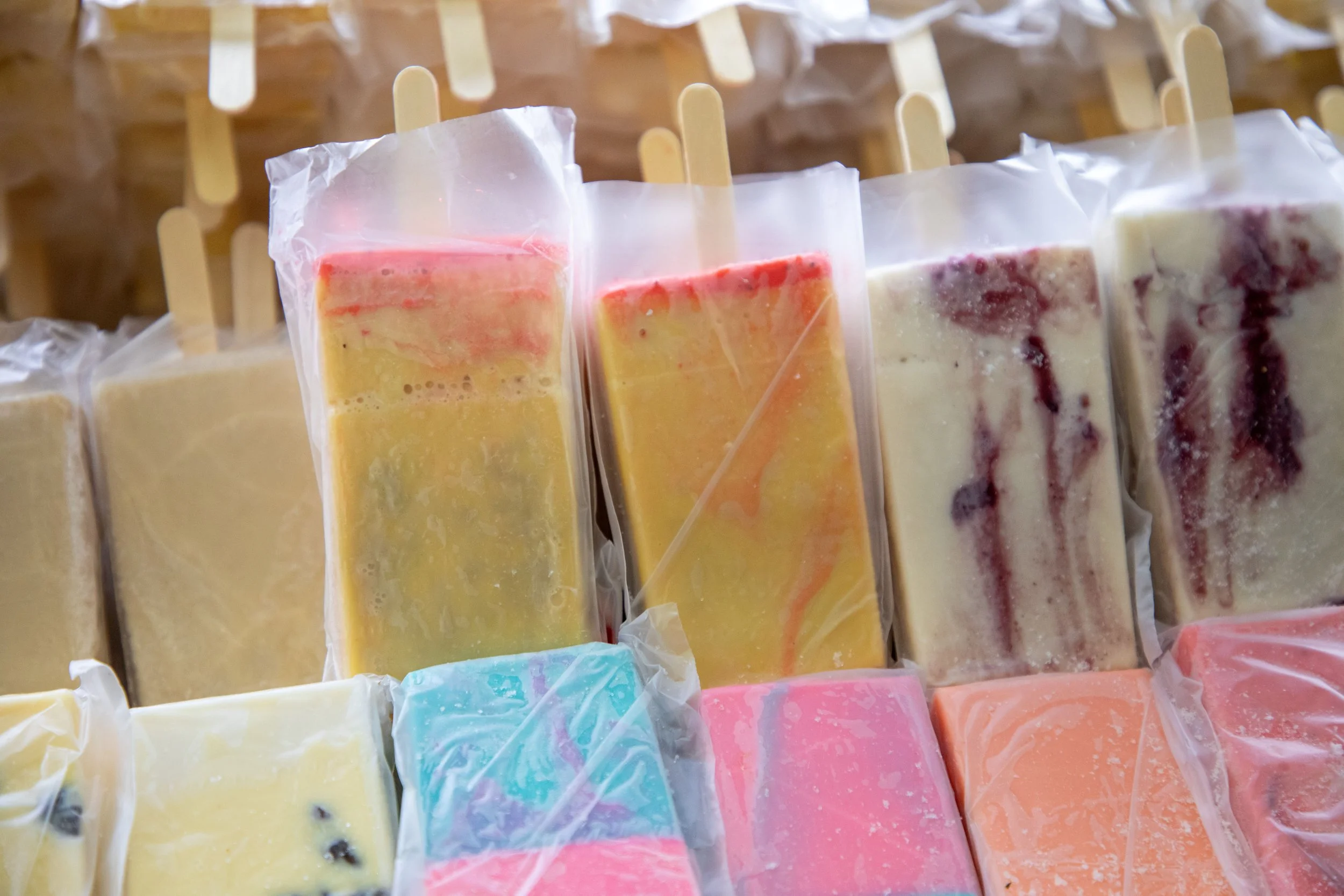 Colorful handmade soap bars arranged on a display shelf, with each soap package wrapped in clear plastic and a wooden stick inserted into each.