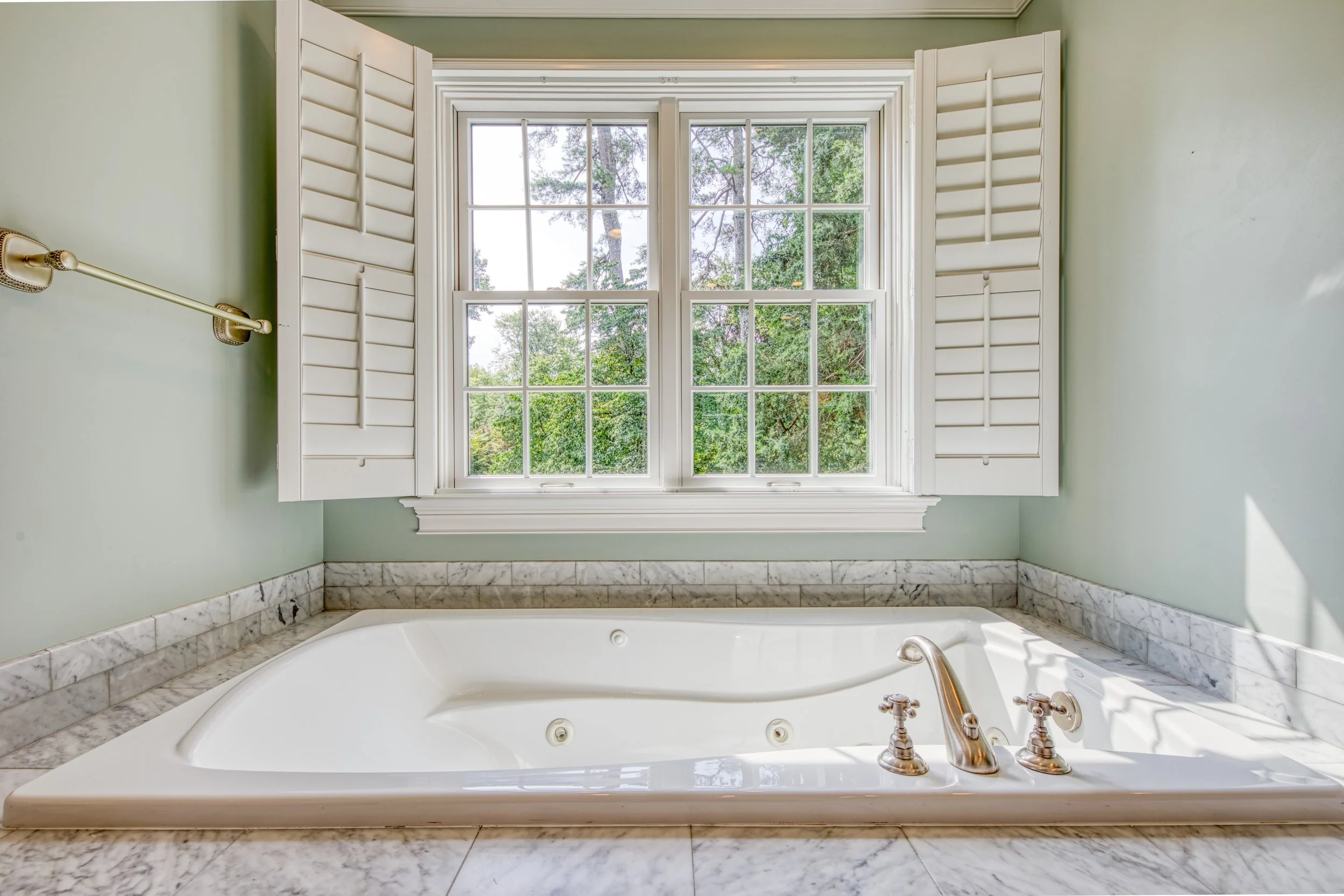 Bathroom with a large window, white plantation shutters, and a whirlpool bathtub with marble surround.