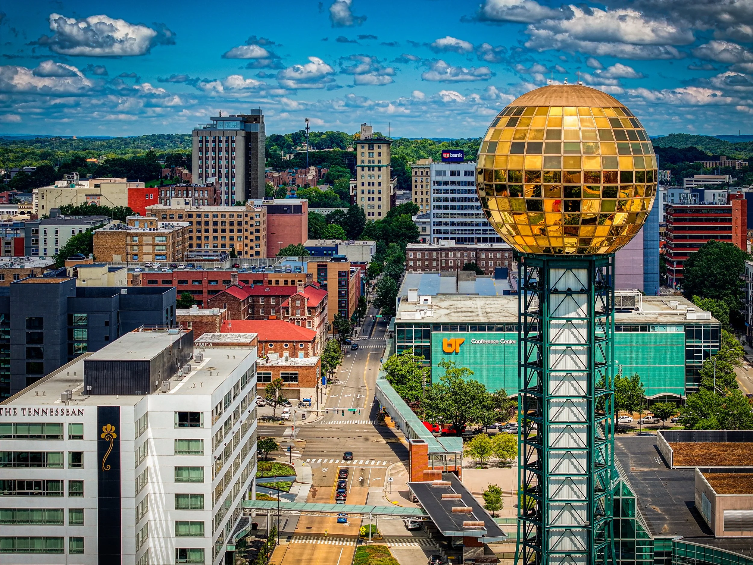 City skyline featuring a large gold and glass spherical structure on a tower, surrounded by various buildings, streets, and trees under a partly cloudy sky.