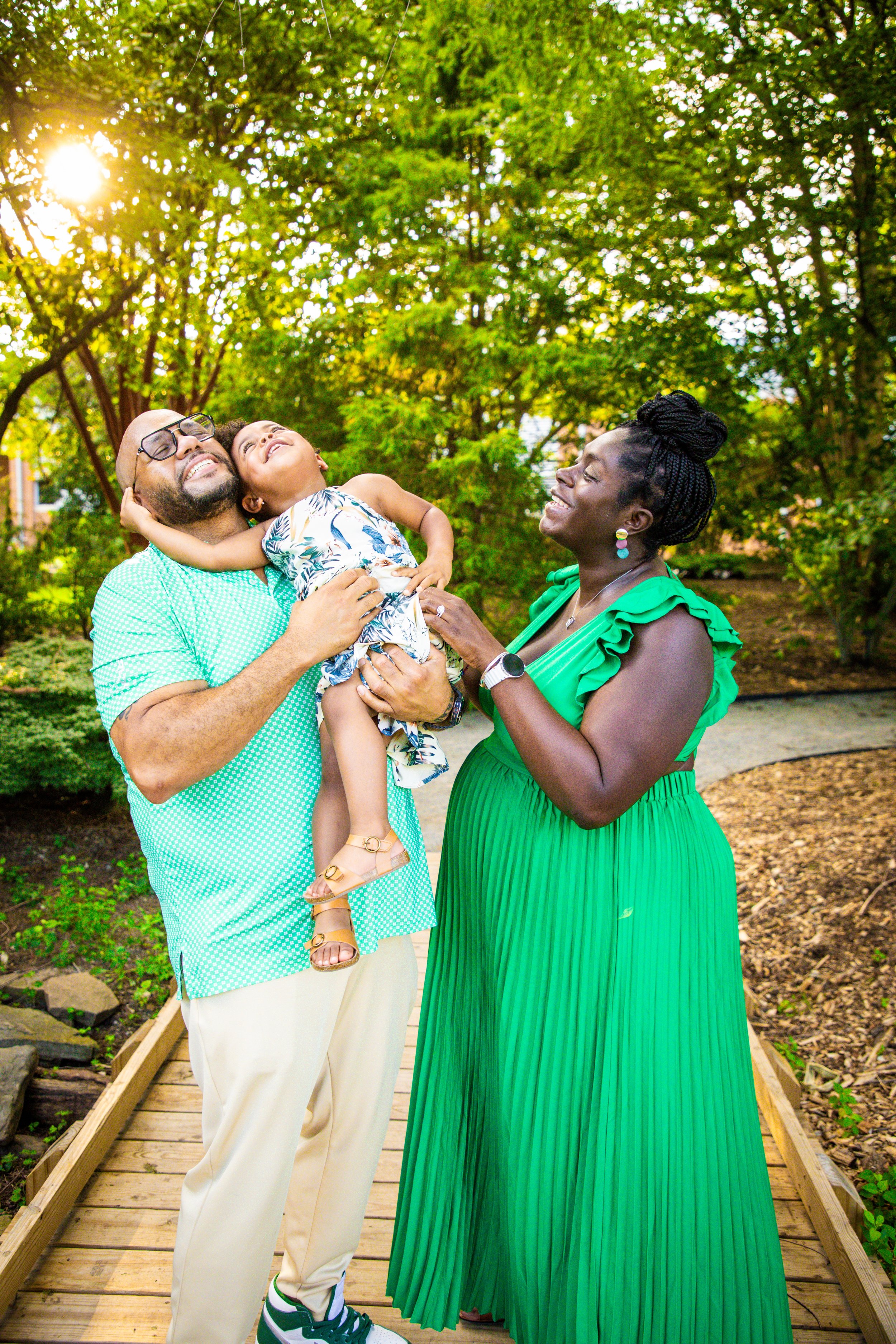 A family enjoying a moment outdoors under the sunlight, standing on a wooden pathway surrounded by green trees and plants.
