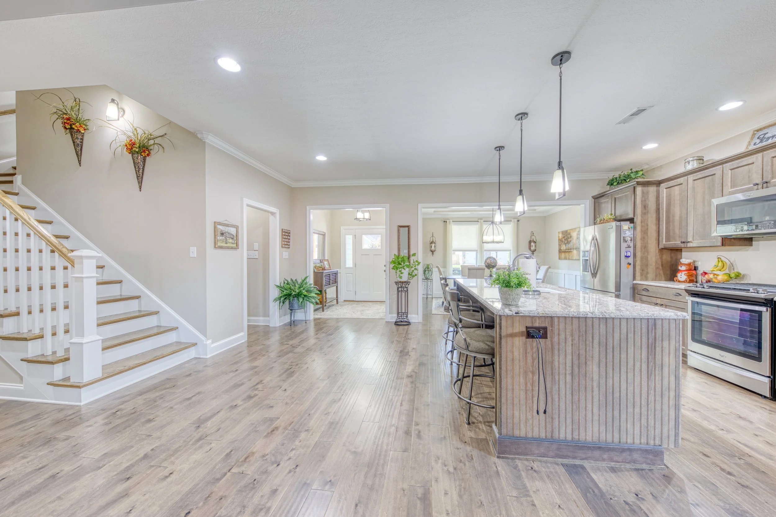 Open-concept kitchen and living area with wooden flooring, large kitchen island, pendant lighting, and decorative wall sconces, with stairs on the left and a dining area in the background.