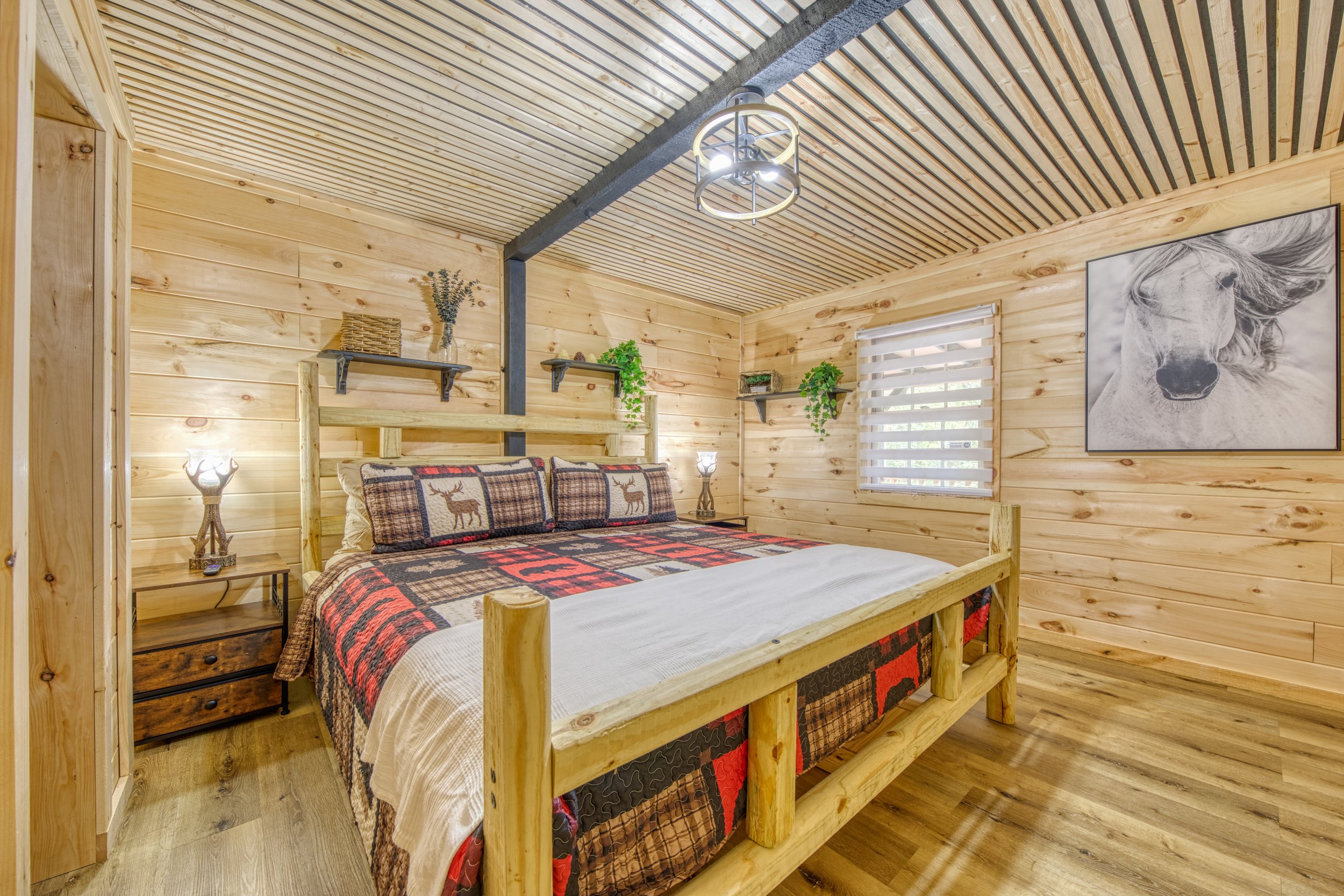 A cozy bedroom with wooden walls and ceiling, a bed with a rustic log frame, plaid bedspread, and deer-themed pillows, wooden nightstand with a lamp, and a large black and white horse portrait on the wall.