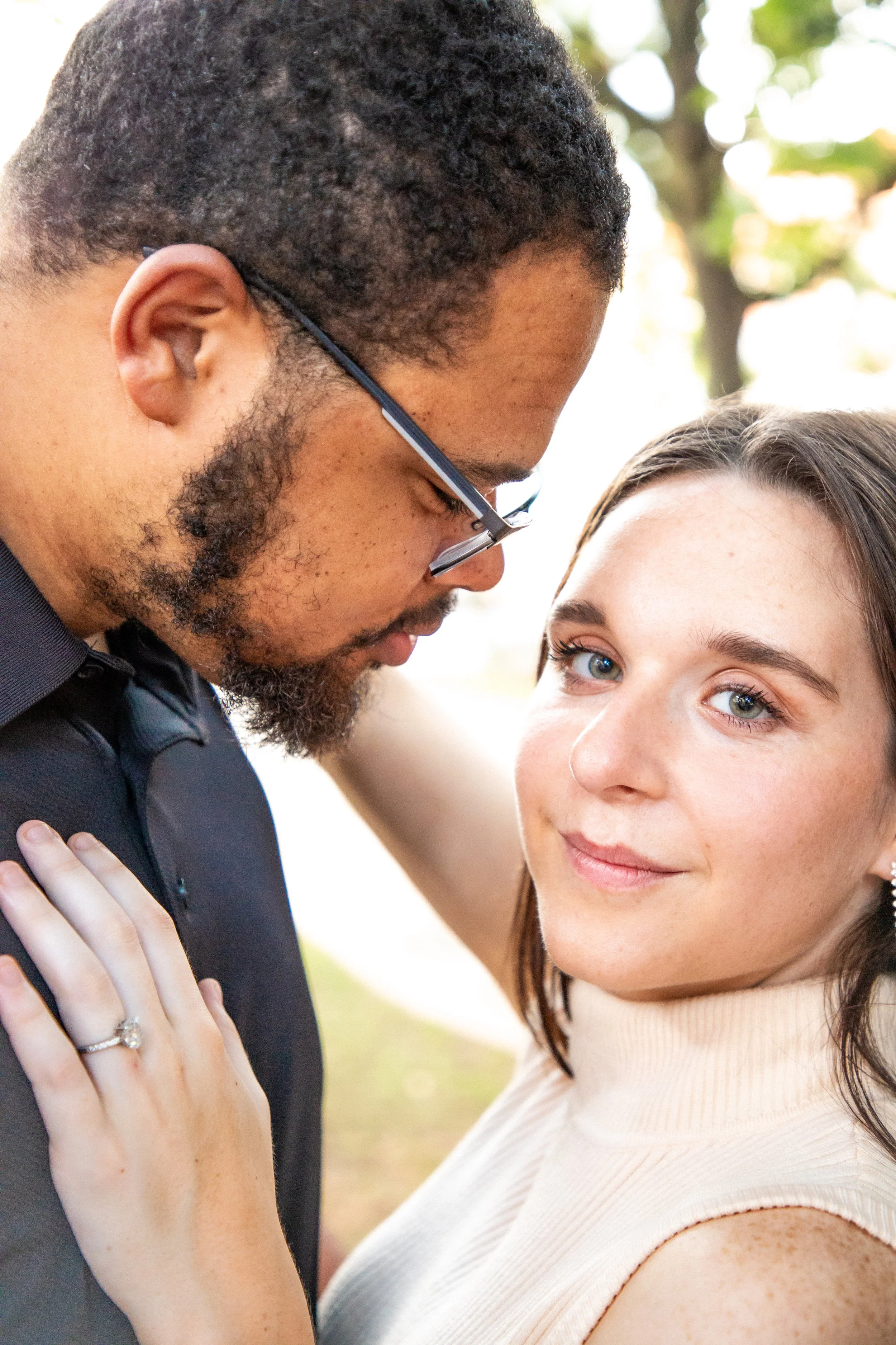 Close-up of a couple, with the man whispering into the woman's ear, outdoors with blurred greenery in the background.