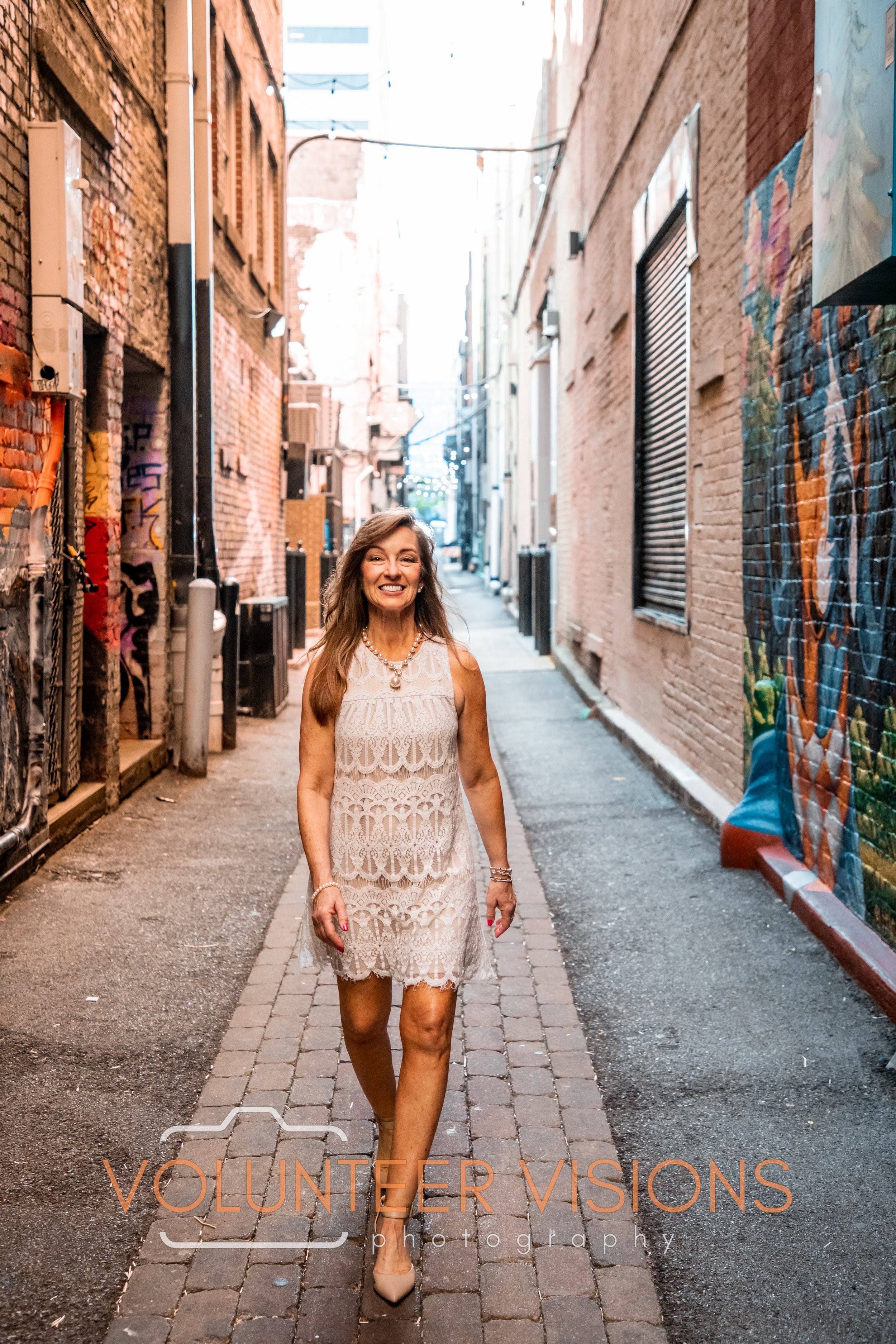 A woman in a white lace dress walking down a brick alleyway, smiling, with colorful graffiti on the right wall and buildings on each side.