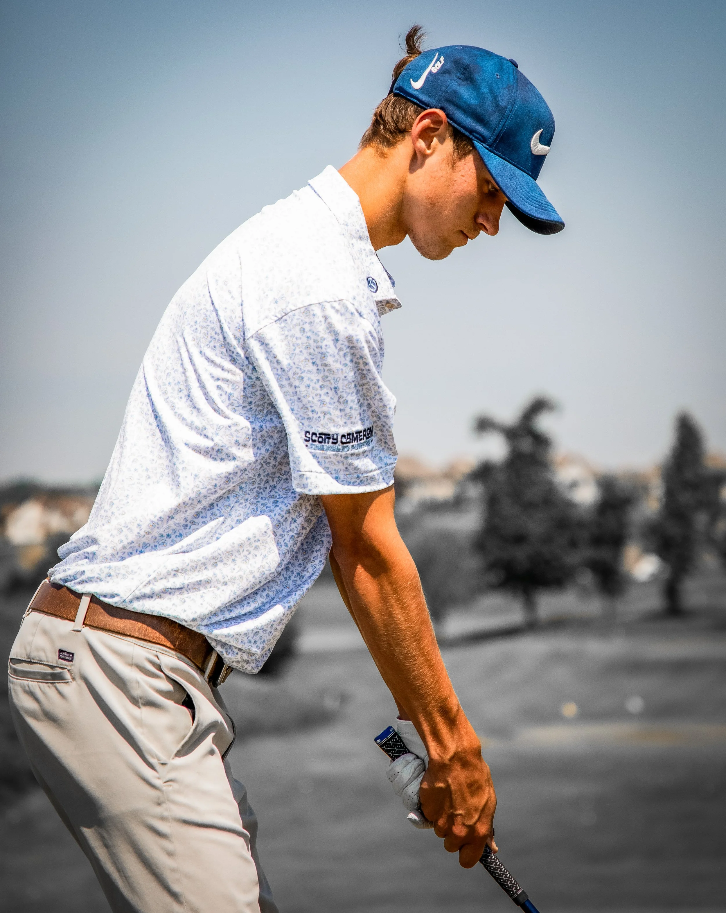 A male golfer is on a golf course, wearing a blue cap, white shirt, and beige pants, preparing to take a shot.