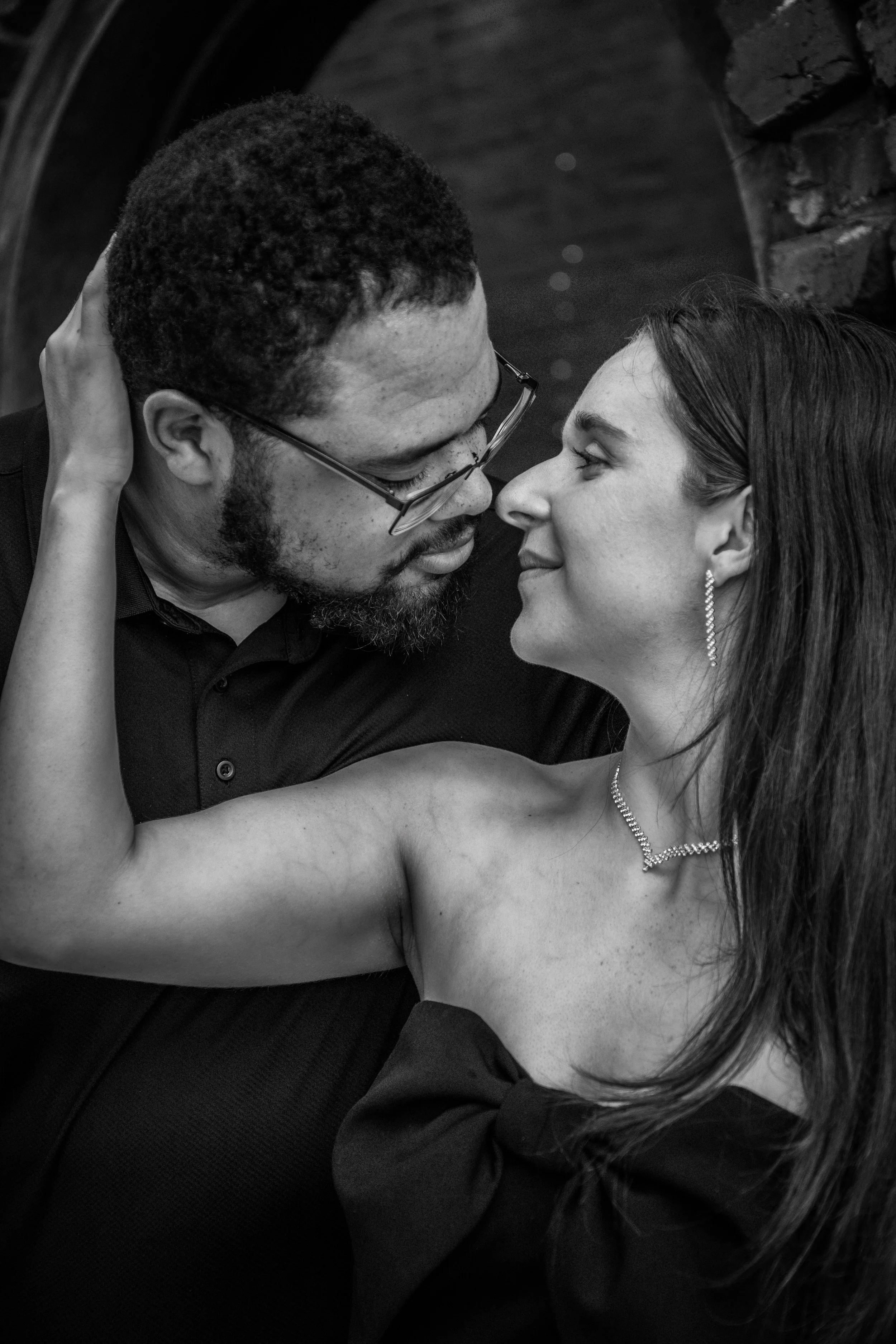 A black and white close-up of a couple in an intimate moment, with the man holding the woman around her shoulders and the woman looking into the man's eyes, both smiling.