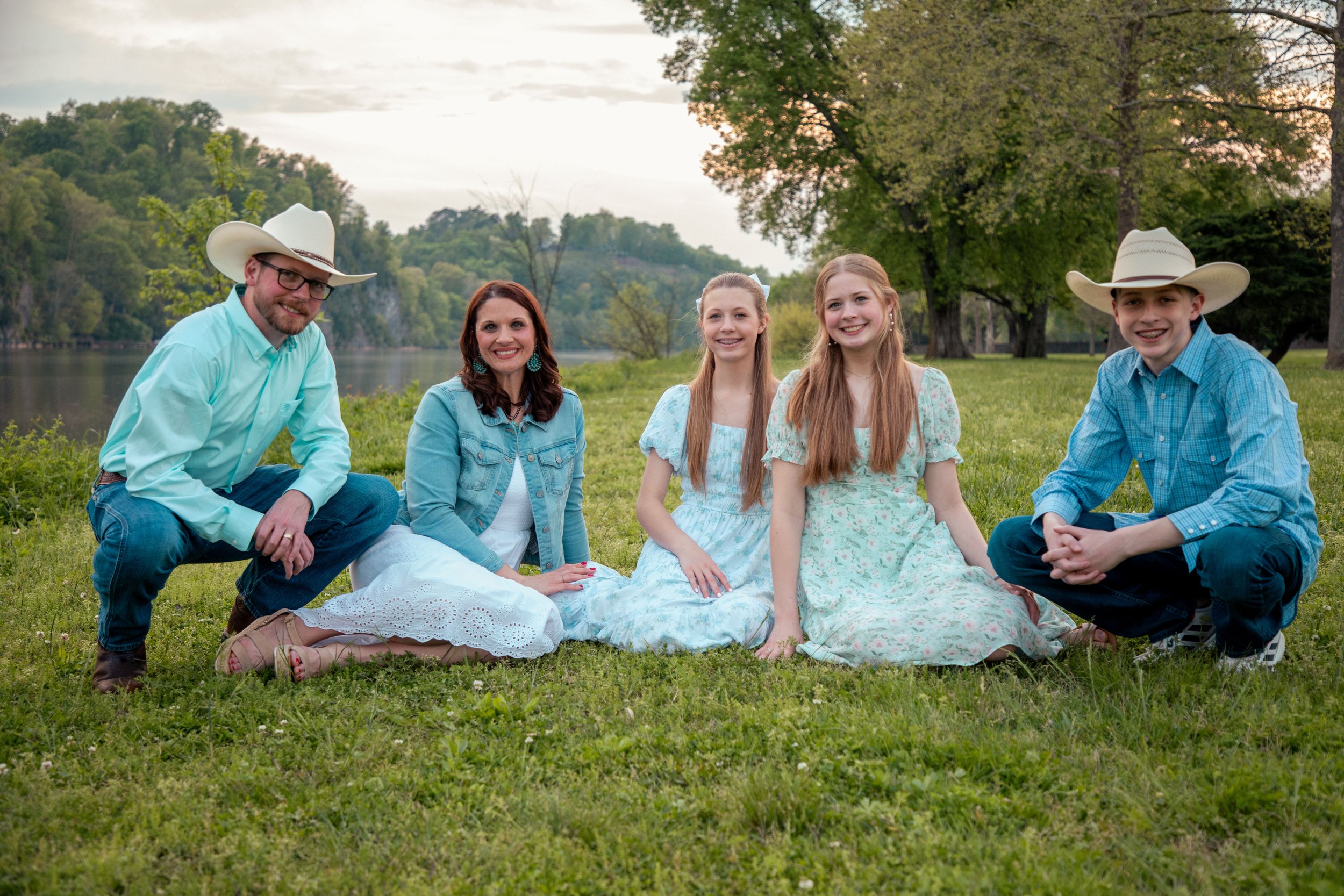 A family of five sitting on the grass near a river with lush green trees and hills in the background. Two young women and two young men wearing cowboy hats and casual clothes, and an older woman wearing earrings and a denim jacket.