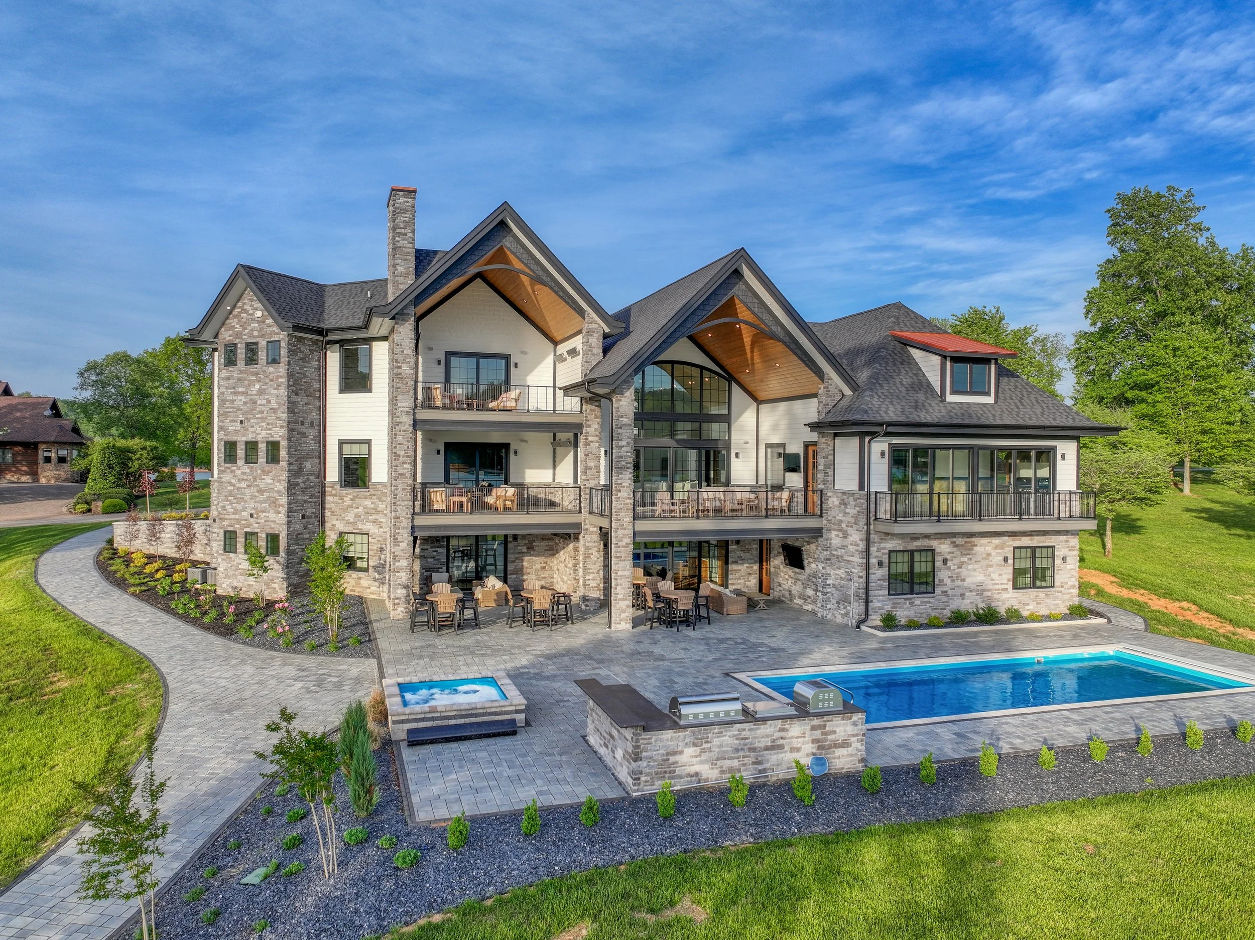 Large modern house with stone accents, multiple balconies, and a backyard with a swimming pool and hot tub, surrounded by green lawn and trees under a blue sky.