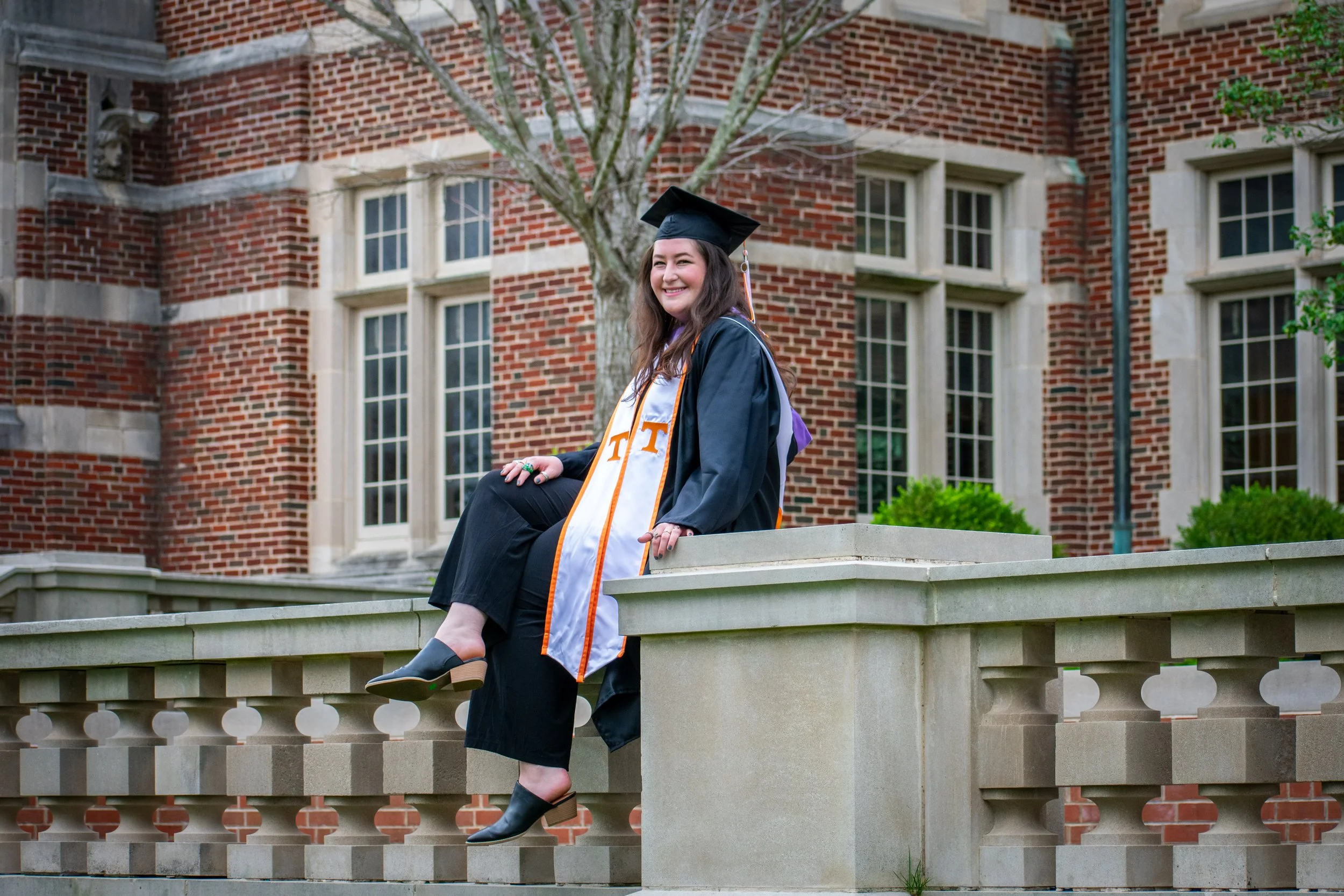 A young woman in a graduation gown and cap sitting on a stone ledge outside a red brick building, smiling and looking to the side.