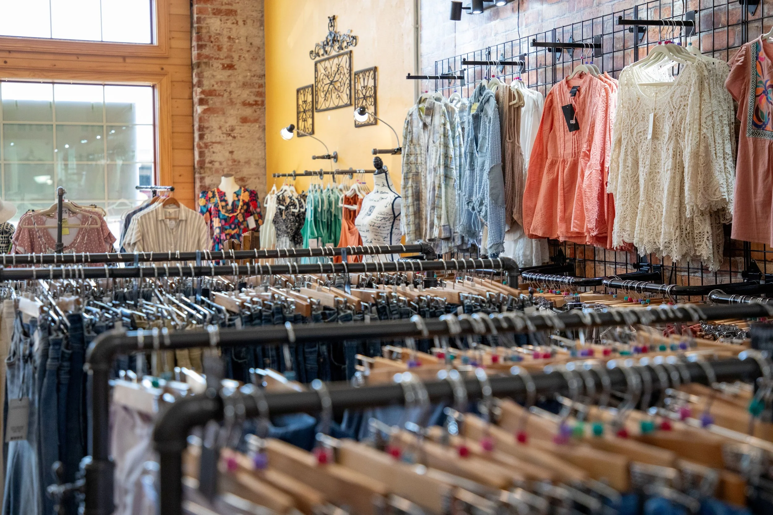Clothing store interior with racks of women’s dresses, blouses, and shirts, against a brick wall and large windows.