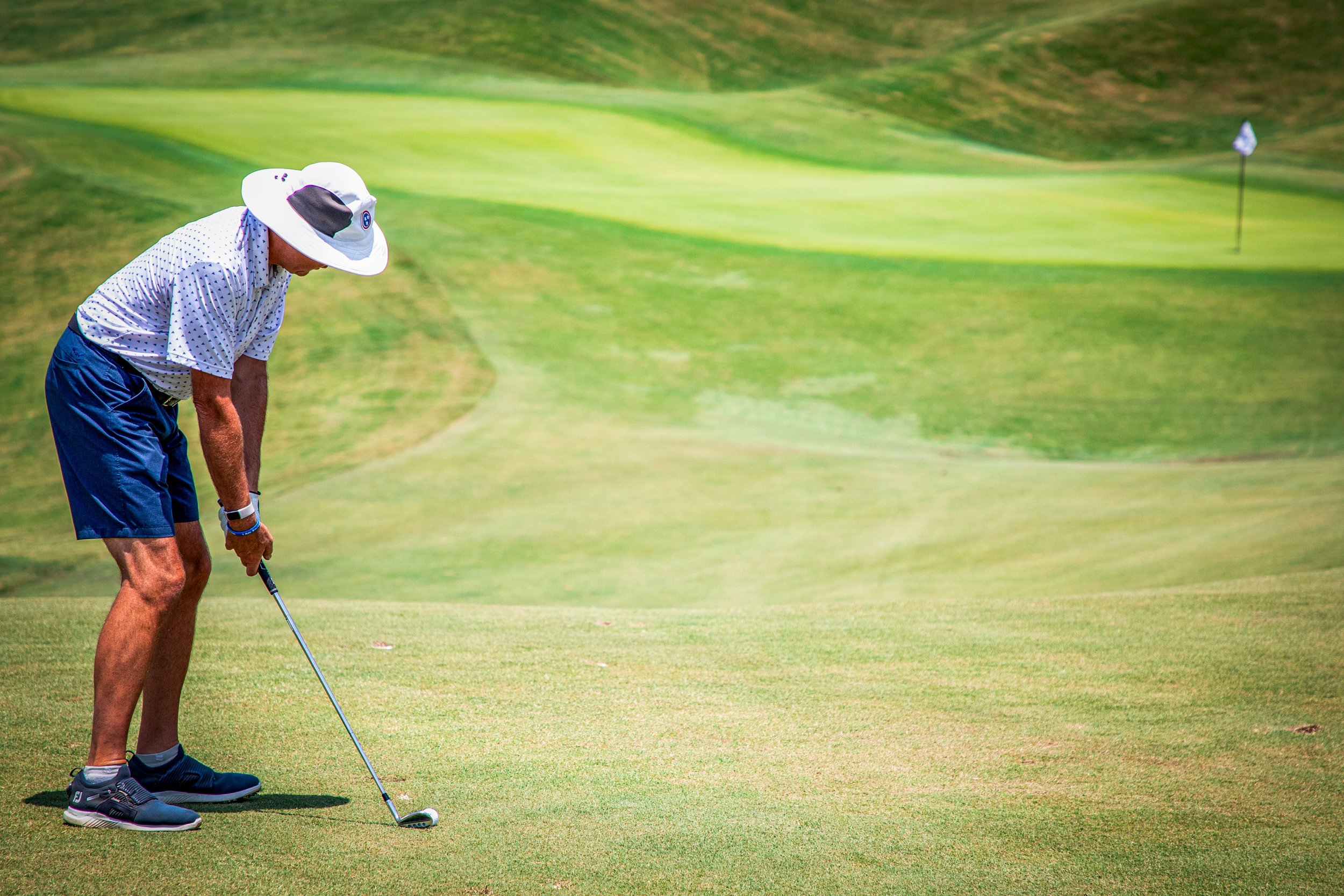 A man wearing a white hat, a white shirt with blue polka dots, blue shorts, and gray sneakers is preparing to putt on a golf course. He is standing on the green, focused on the golf ball with a golf club in his hands, and a hole with a flag is visibl