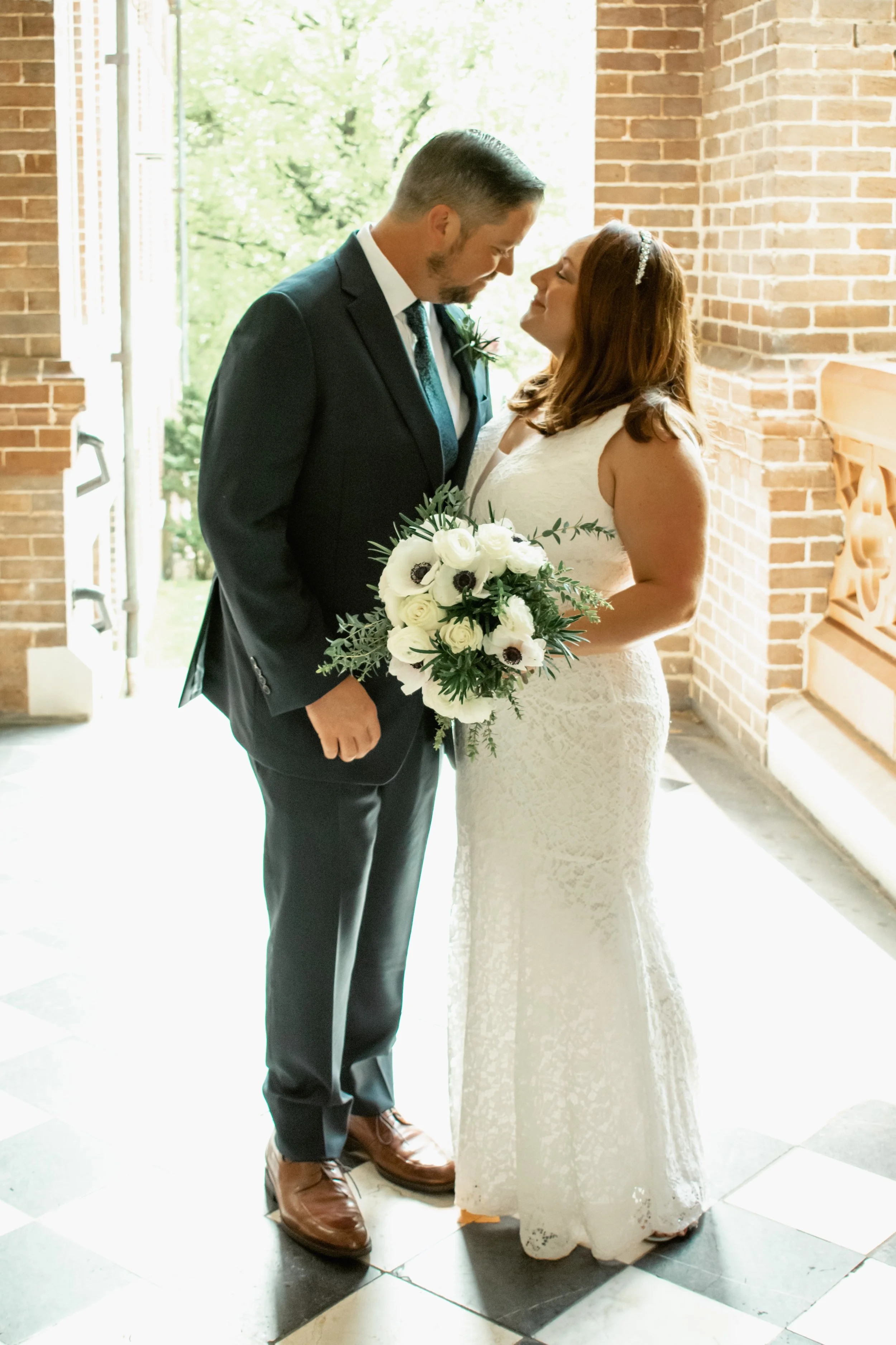 A bride and groom standing close together, touching foreheads, during a wedding ceremony inside a building with brick walls and large windows. The groom is wearing a black suit with a boutonniere, and the bride is in a white lace wedding dress holdin