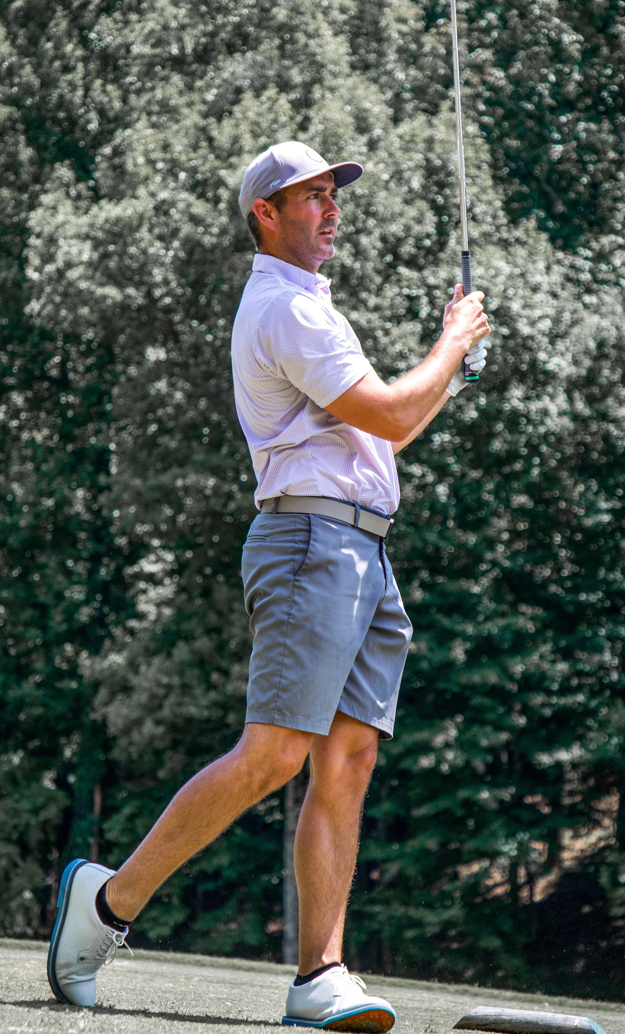 A man playing golf, holding a golf club, wearing a gray cap, light-colored shirt, gray shorts, and white golf shoes, standing on a golf course with trees in the background.