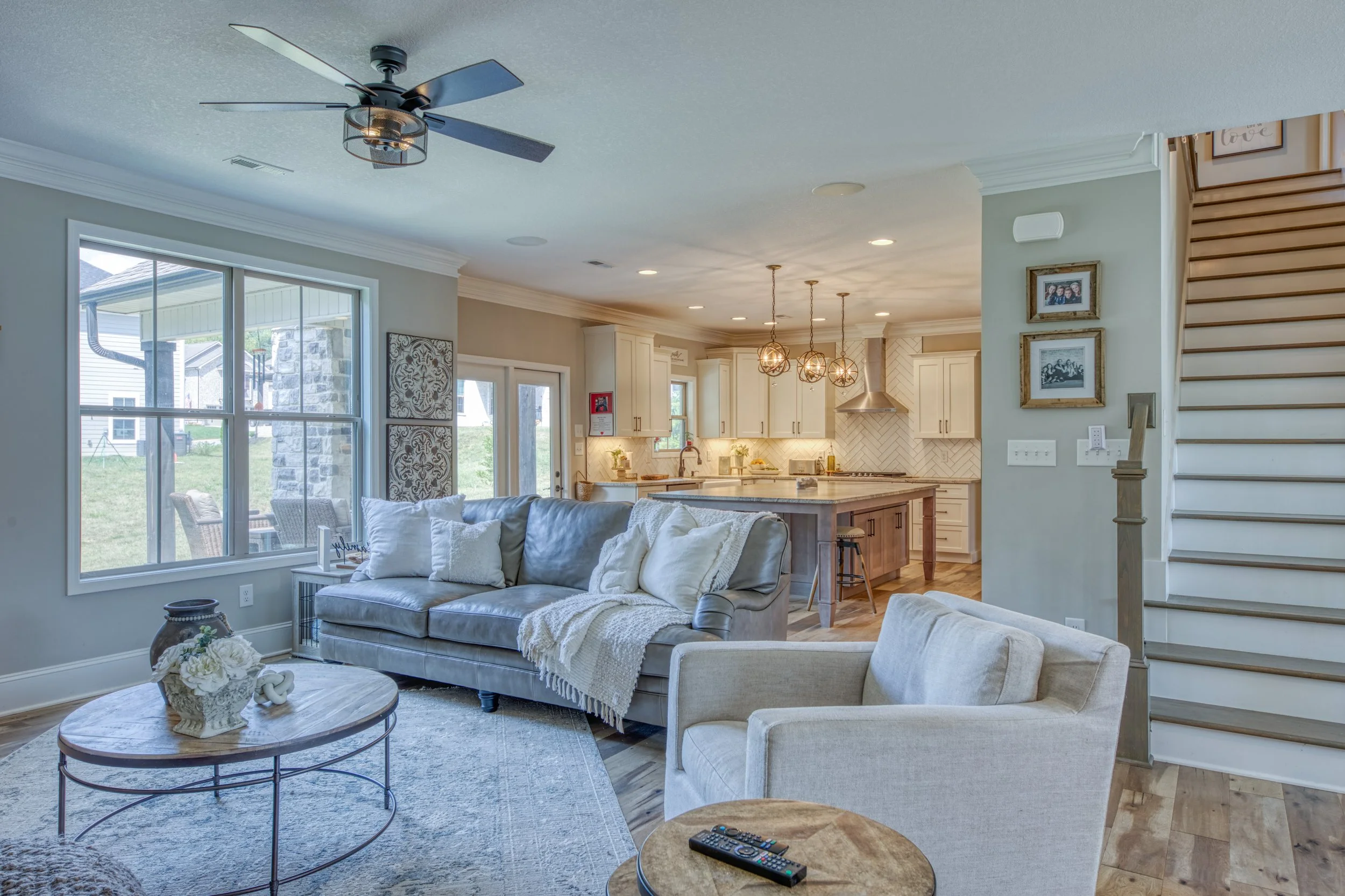 Living room with large window, beige carpet, gray sofa with white pillows, armchair, wooden coffee table with flower bouquet, open kitchen area with white cabinets, kitchen island, hanging pendant lights, staircase with wooden steps, family photos on