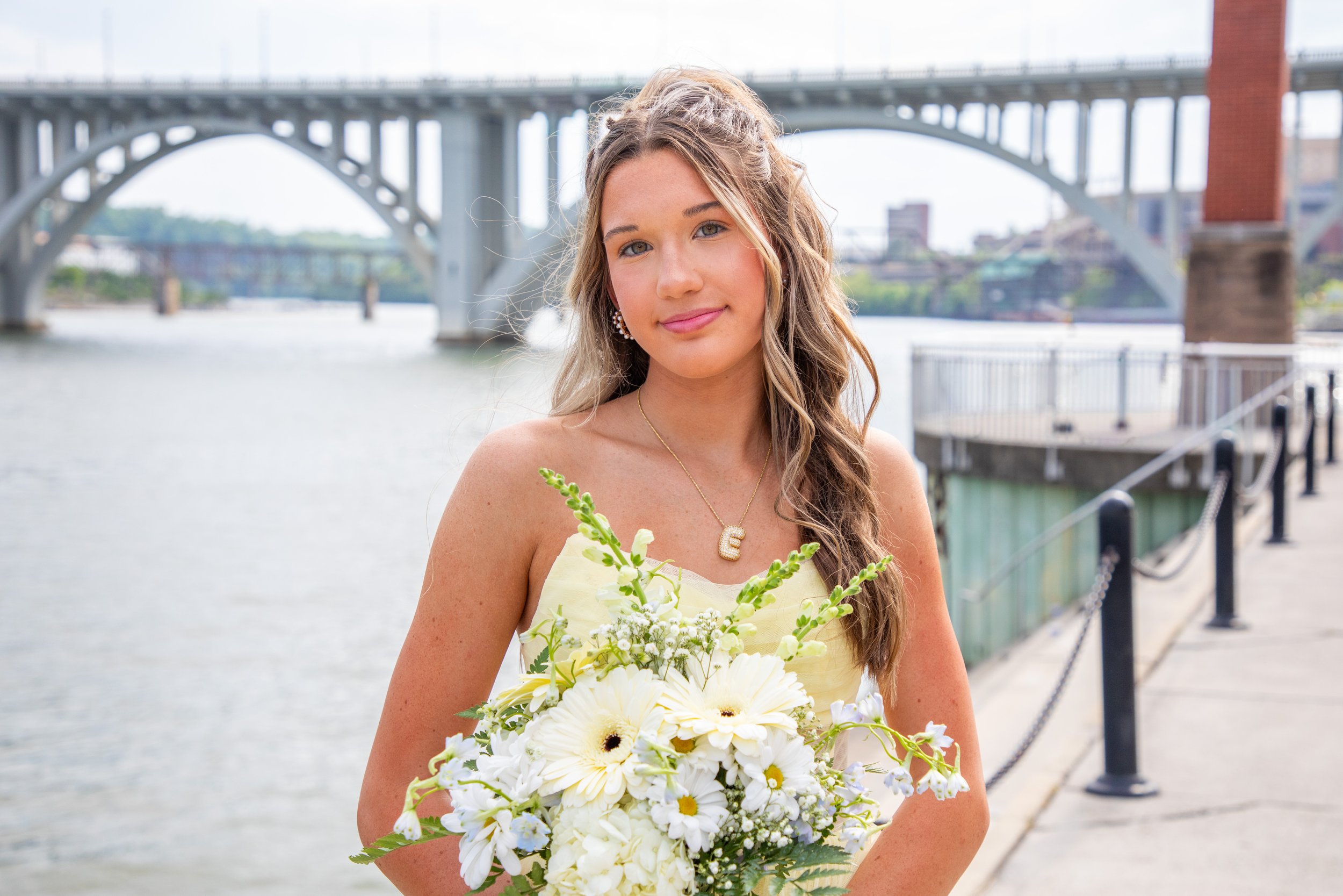 A young woman with long wavy hair and a light complexion holding a bouquet of white flowers, standing near a river with a bridge in the background.