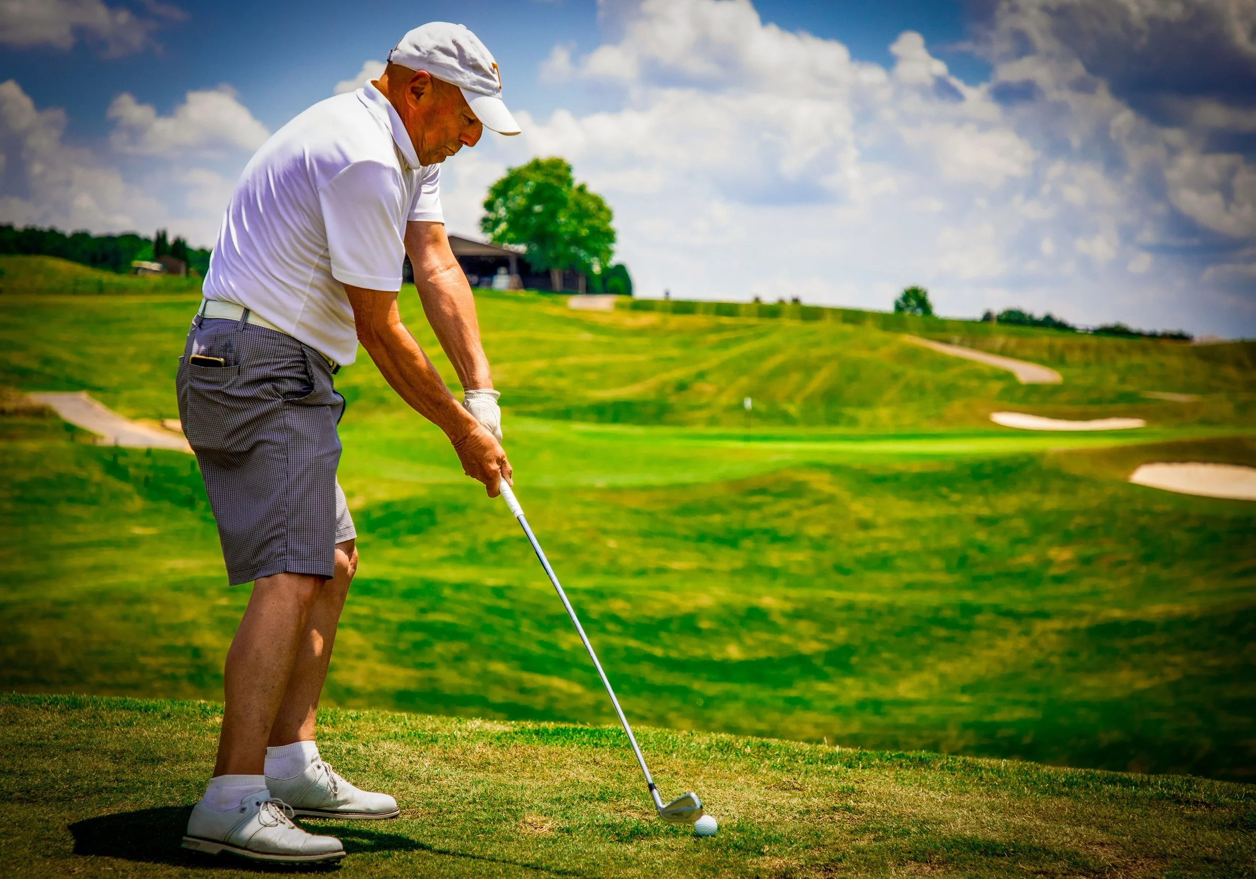 An elderly man wearing a white cap, white golf shirt, and checkered shorts, is preparing to hit a golf ball on a lush green golf course under a partly cloudy sky.