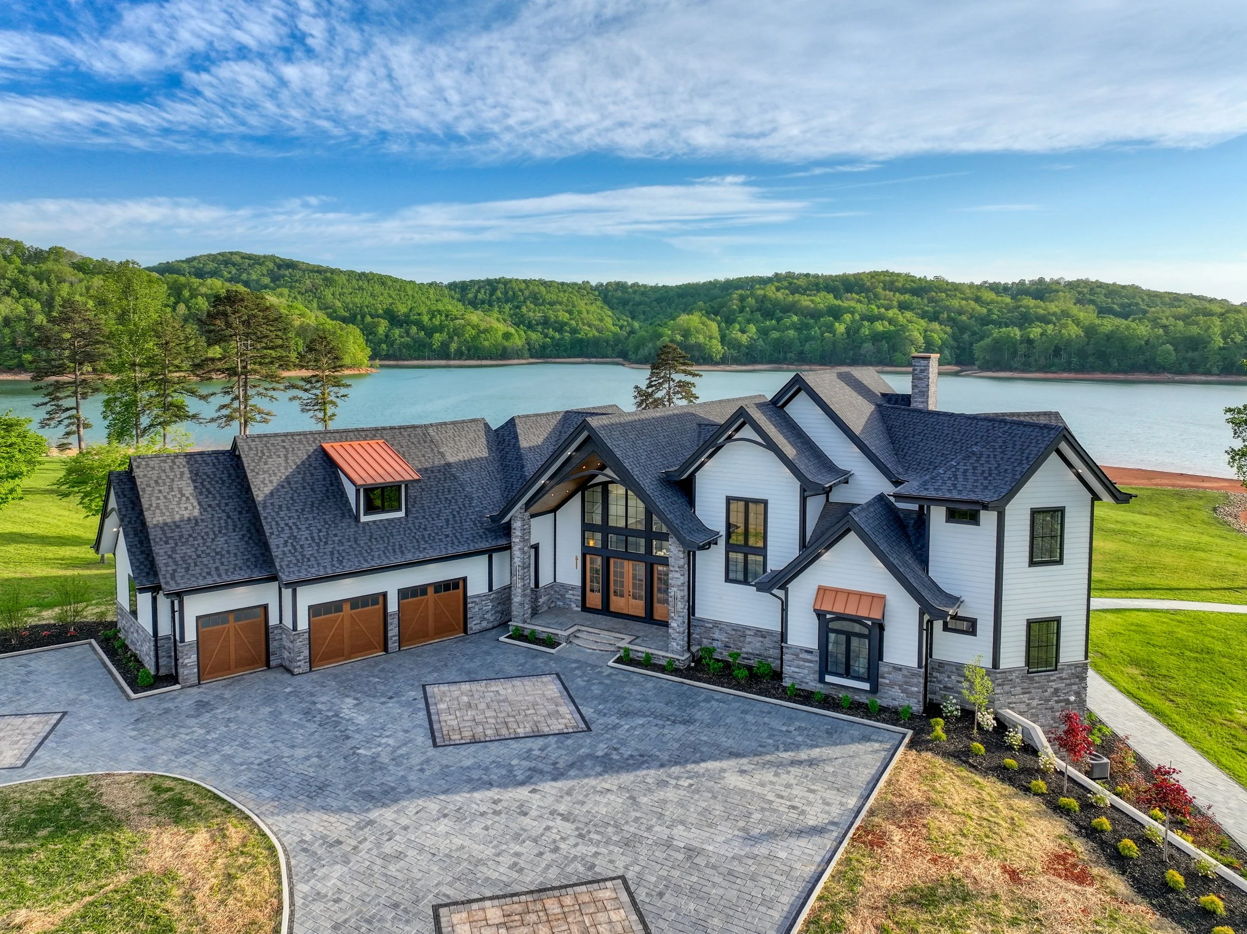 A large modern house with a stone and white exterior, multiple gabled roofs, and a paved driveway surrounded by well-maintained grass and flowerbeds, overlooking a lake with green hills and a partly cloudy blue sky in the background.
