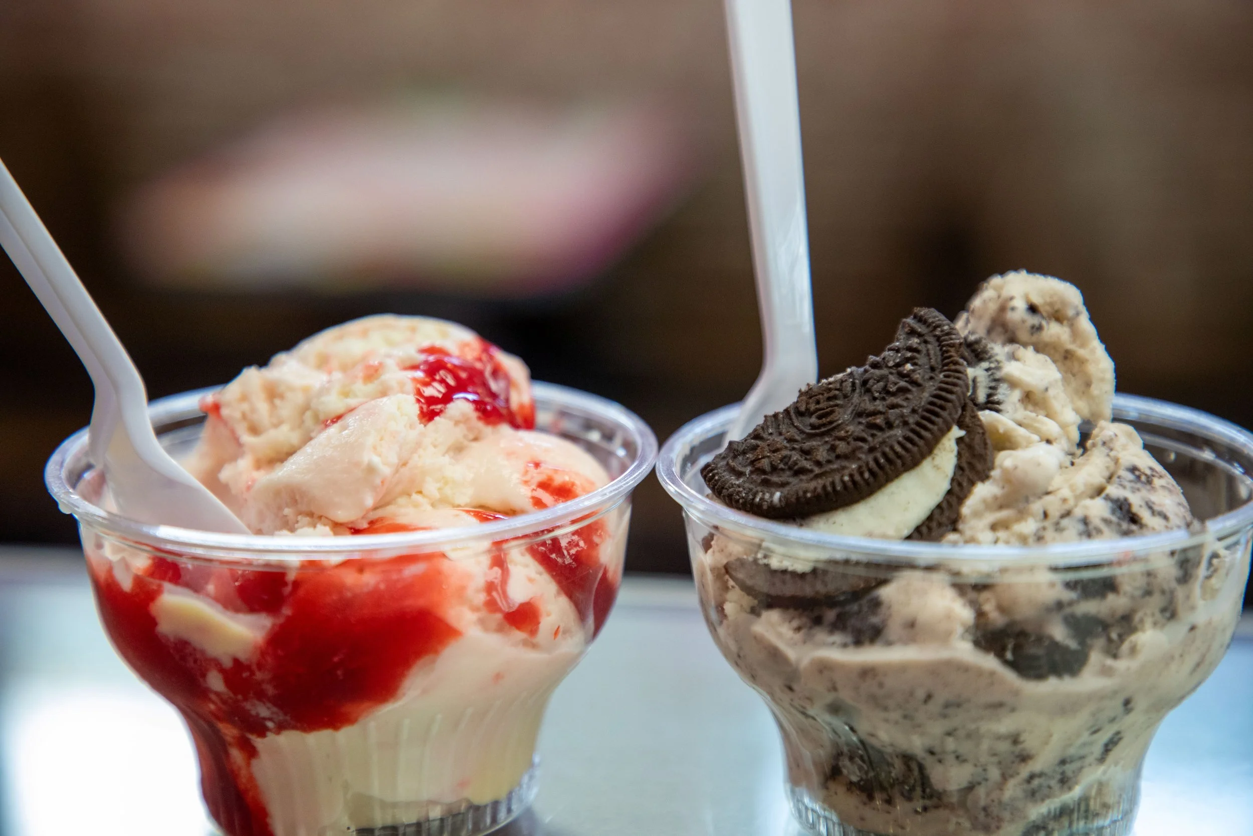Two cups of ice cream with toppings, one with strawberry and the other with cookies and cream, with white spoons inside the cups.