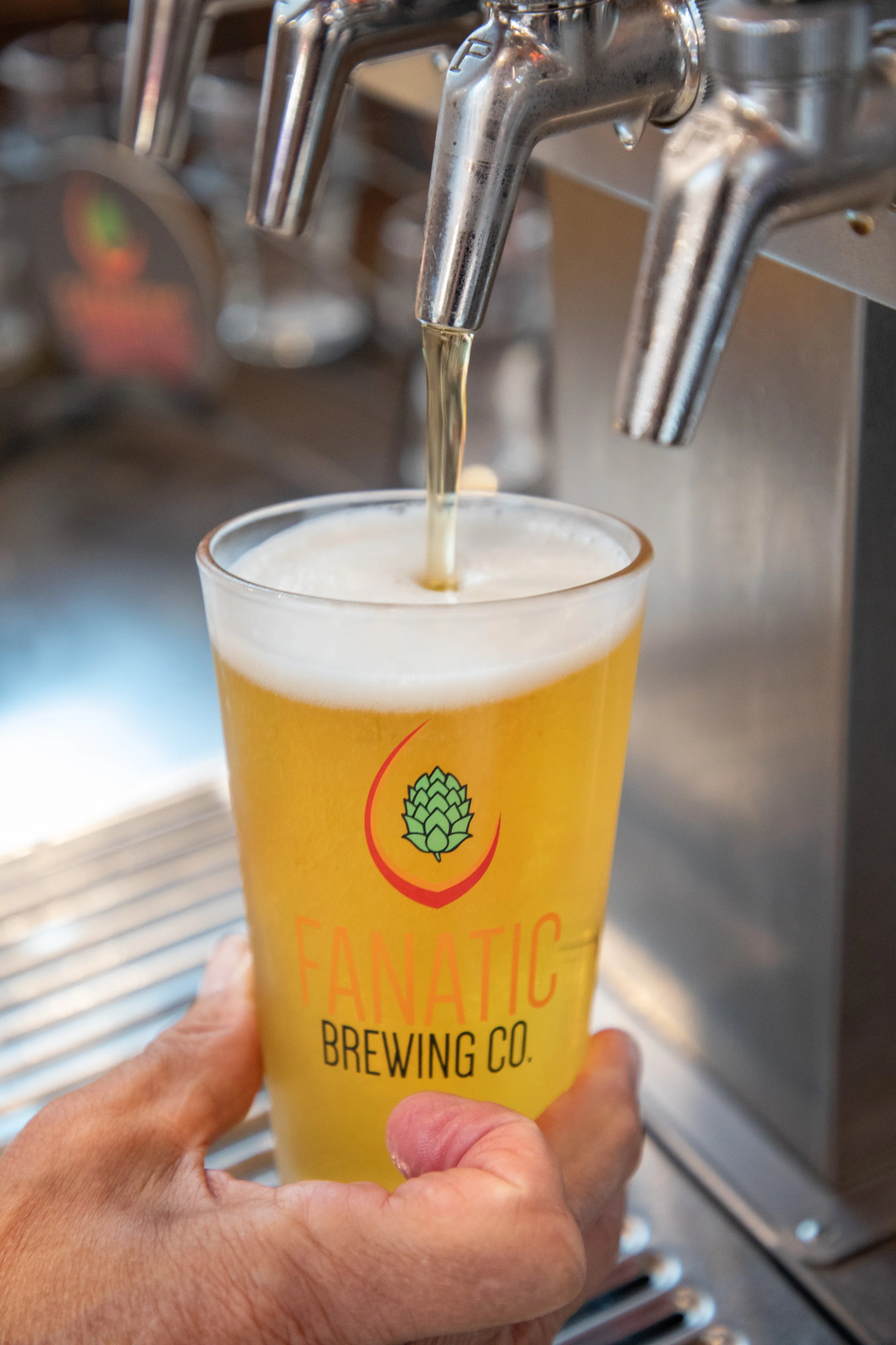 A person holding a tall transparent glass of beer under a tap at a brewery. The glass has the logo of Flatiron Fanatic Brewing Co.
