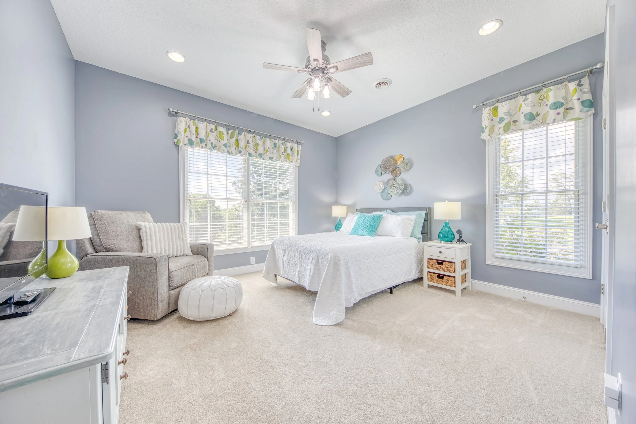 A bright, airy bedroom with large windows, light blue walls, a white bed with turquoise pillows, a gray sofa, a white dresser with a green lamp, and decorative wall art.