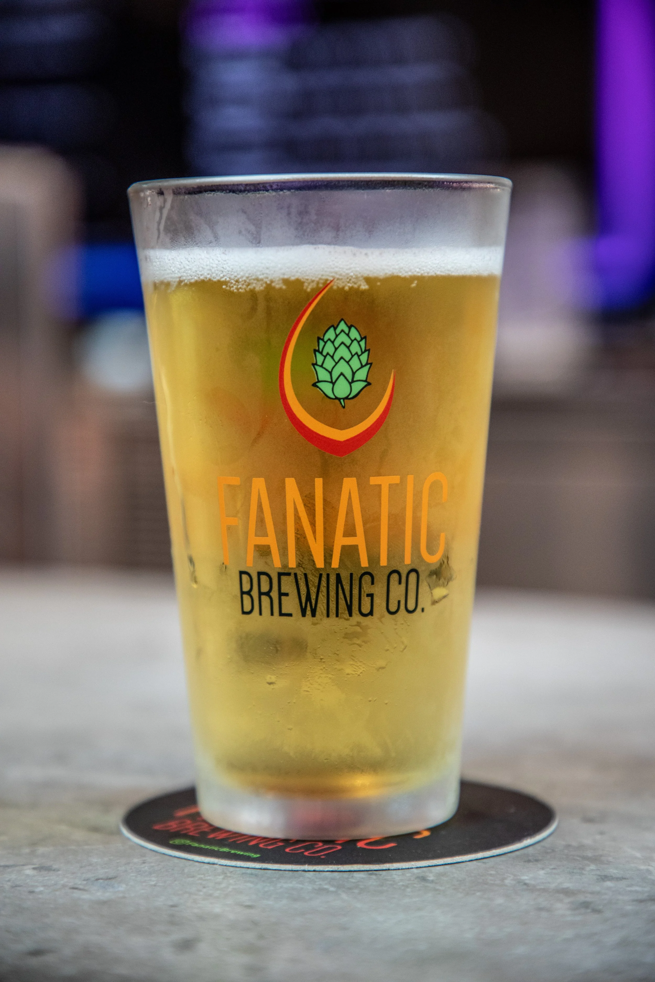 A glass of pale yellow beer with a frothy head, featuring a brewery logo that includes a hop cone and the text 'FANATIC BREWING CO.' The glass is placed on a circular coaster on a concrete surface with a blurred background.
