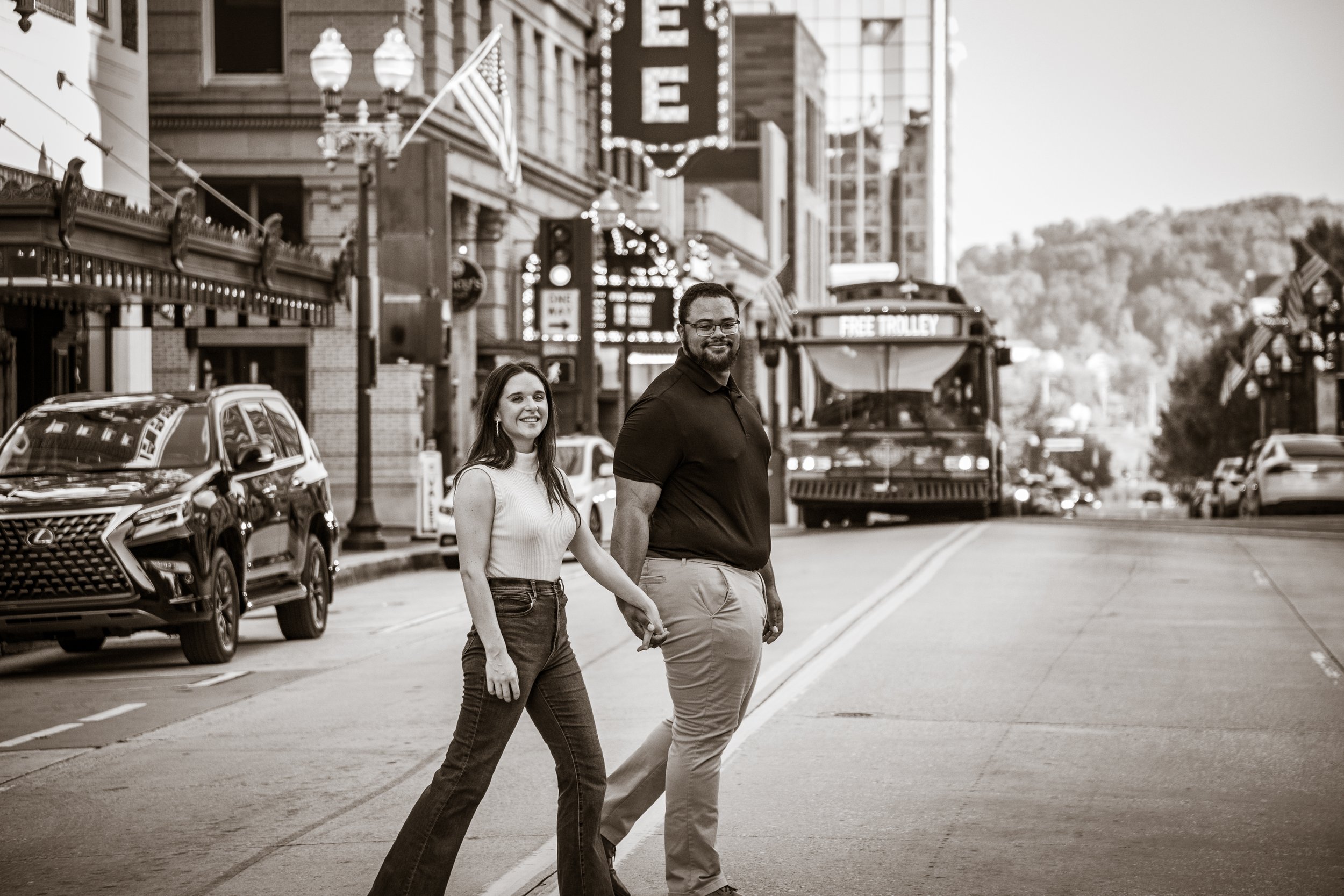 A smiling woman and man holding hands cross the street in a city, with cars parked along the curb and buildings with signs and American flags in the background.