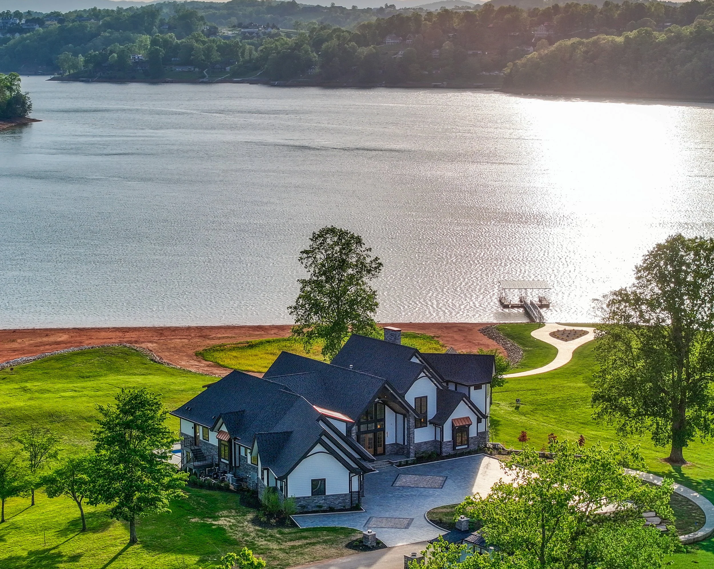 A lake with a small pier and a house by the water, surrounded by green trees and a grassy yard, with houses and a wooded hillside in the background.