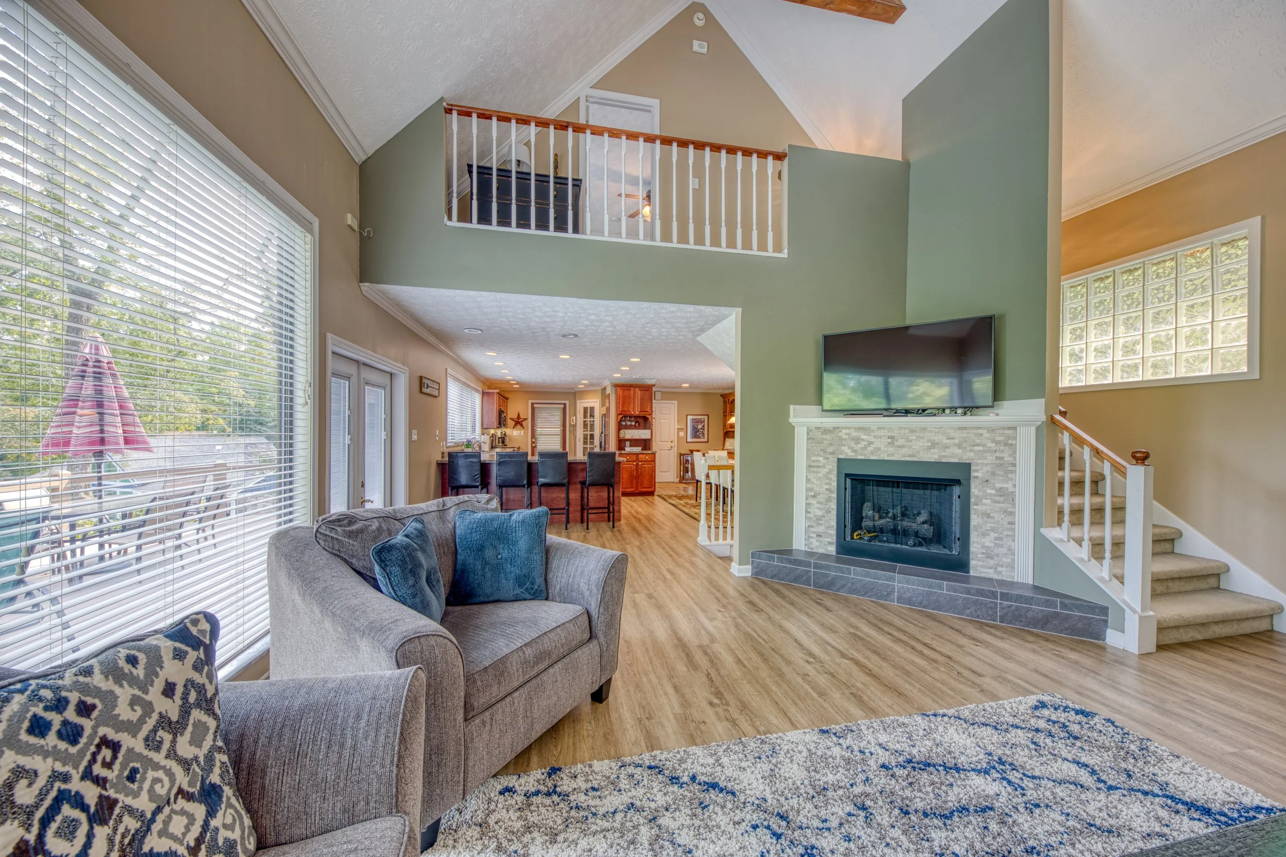 Interior view of a living room with a gray sofa, patterned armchair, hardwood floors, large windows with blinds, and a television above a fireplace, with a view into the kitchen and dining area.