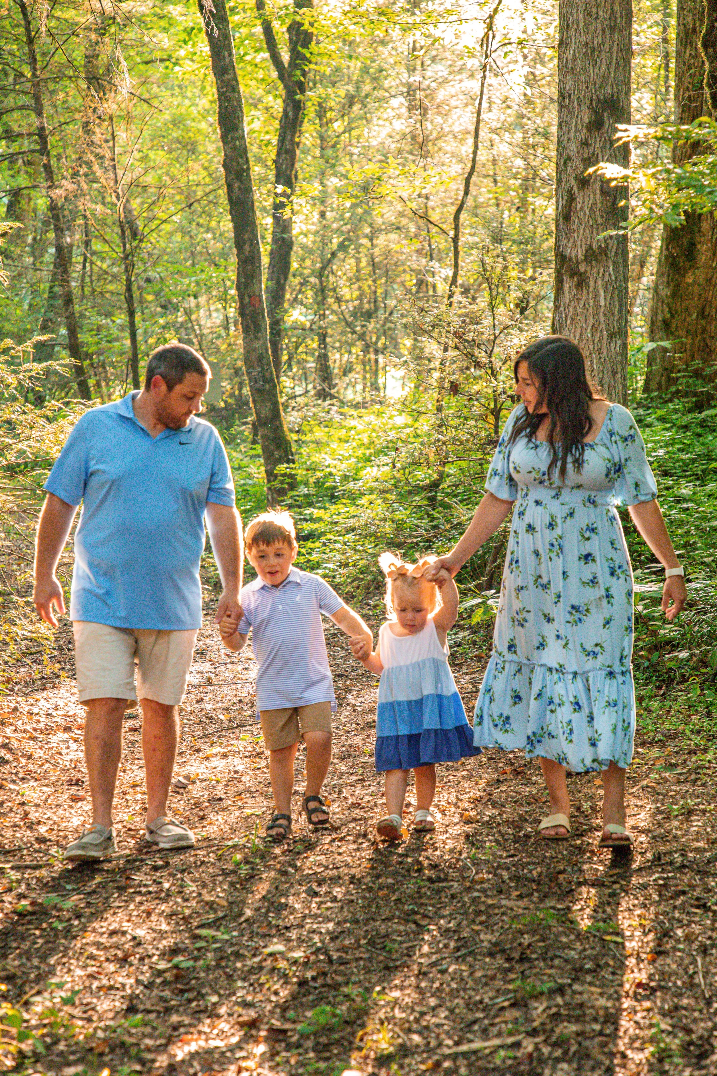 Family walking together on a dirt path in a forest with trees and sunlight filtering through the leaves.