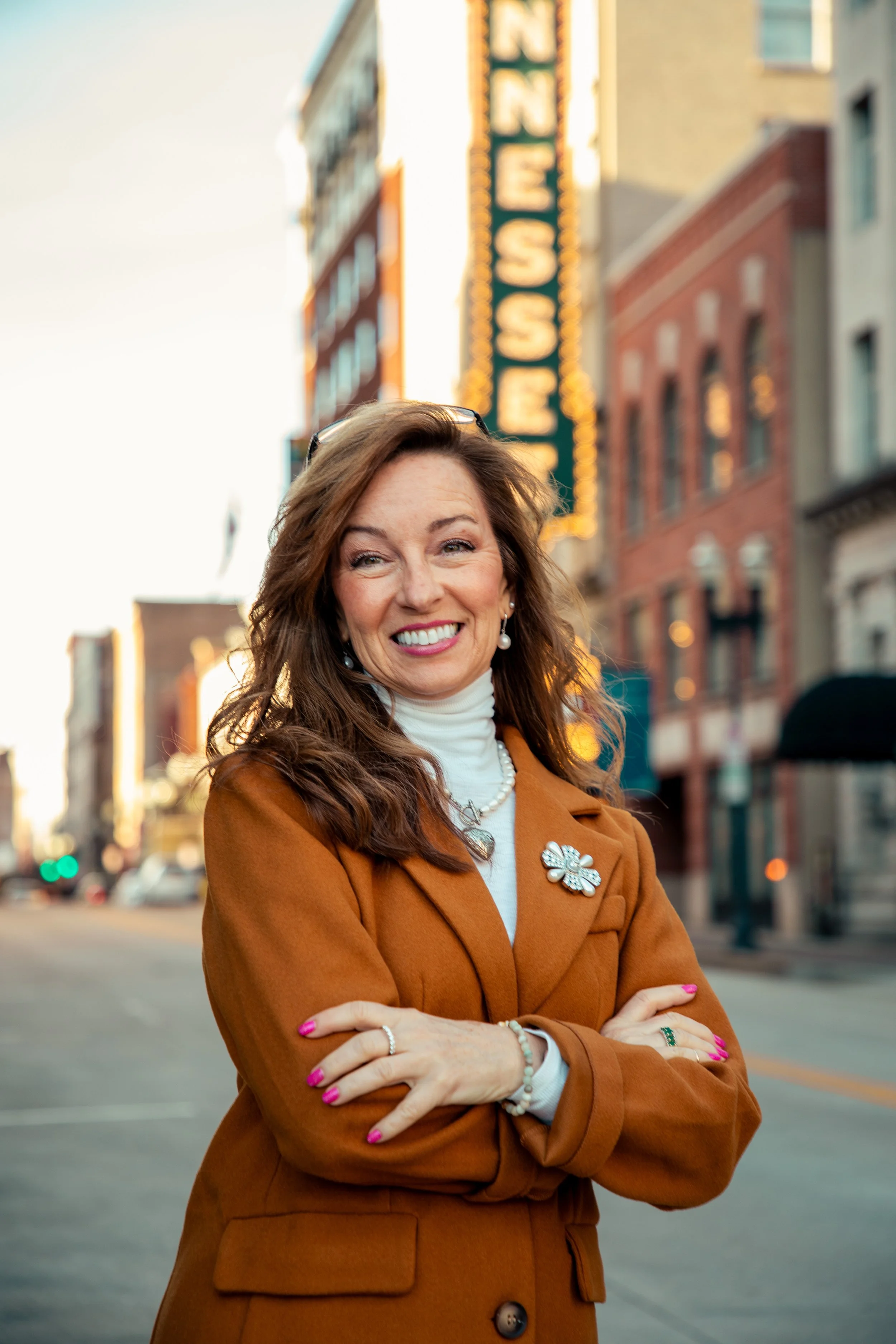 A woman standing on city street with cross-armed pose, smiling, wearing an orange coat, white turtleneck, and jewelry, with buildings and a neon sign in the background.