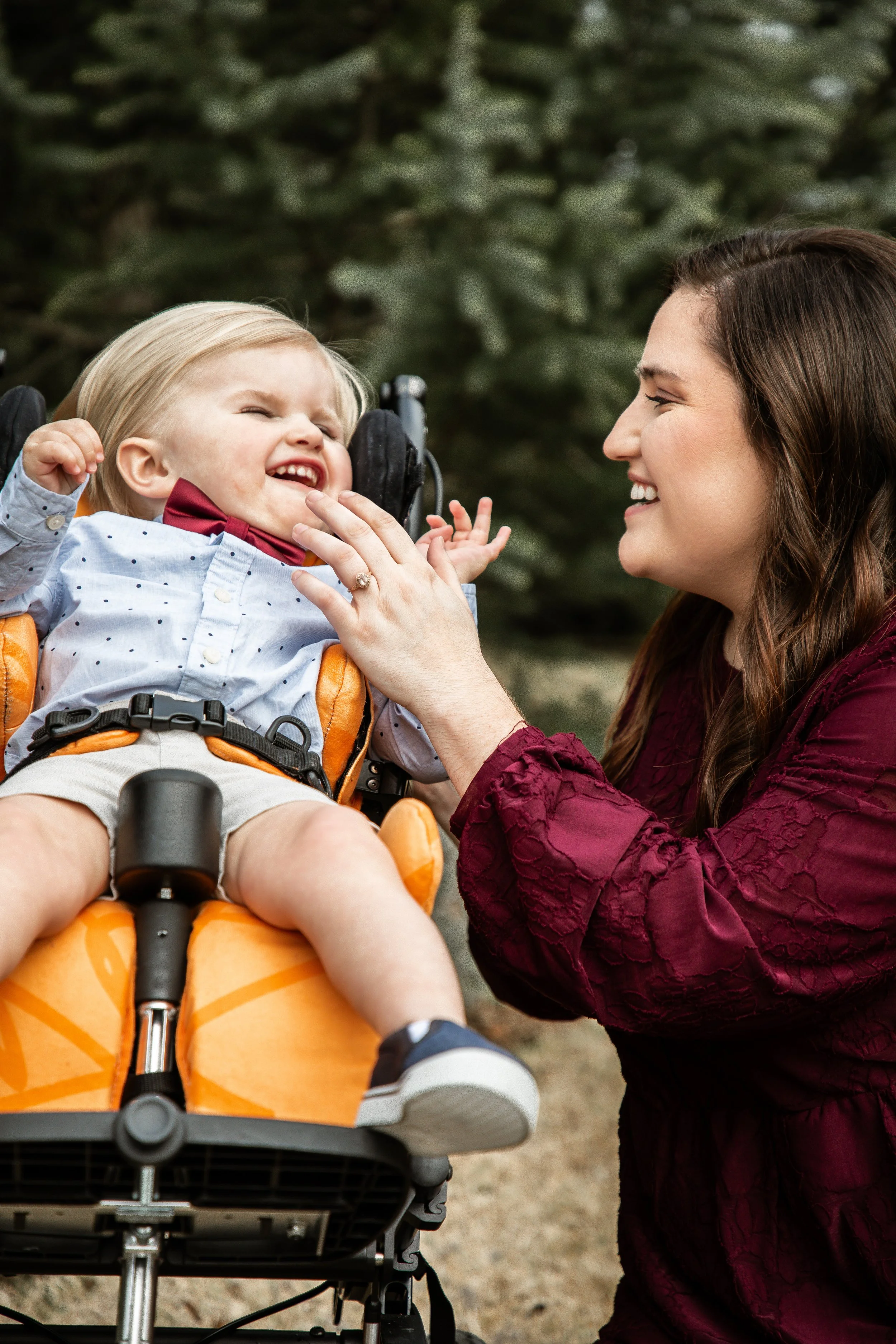 A woman with long brown hair and a smile is interacting with a young boy in a wheelchair. The boy is smiling and wearing a light blue shirt with small dark polka dots, a red bow tie, and beige shorts. The background shows blurred greenery.
