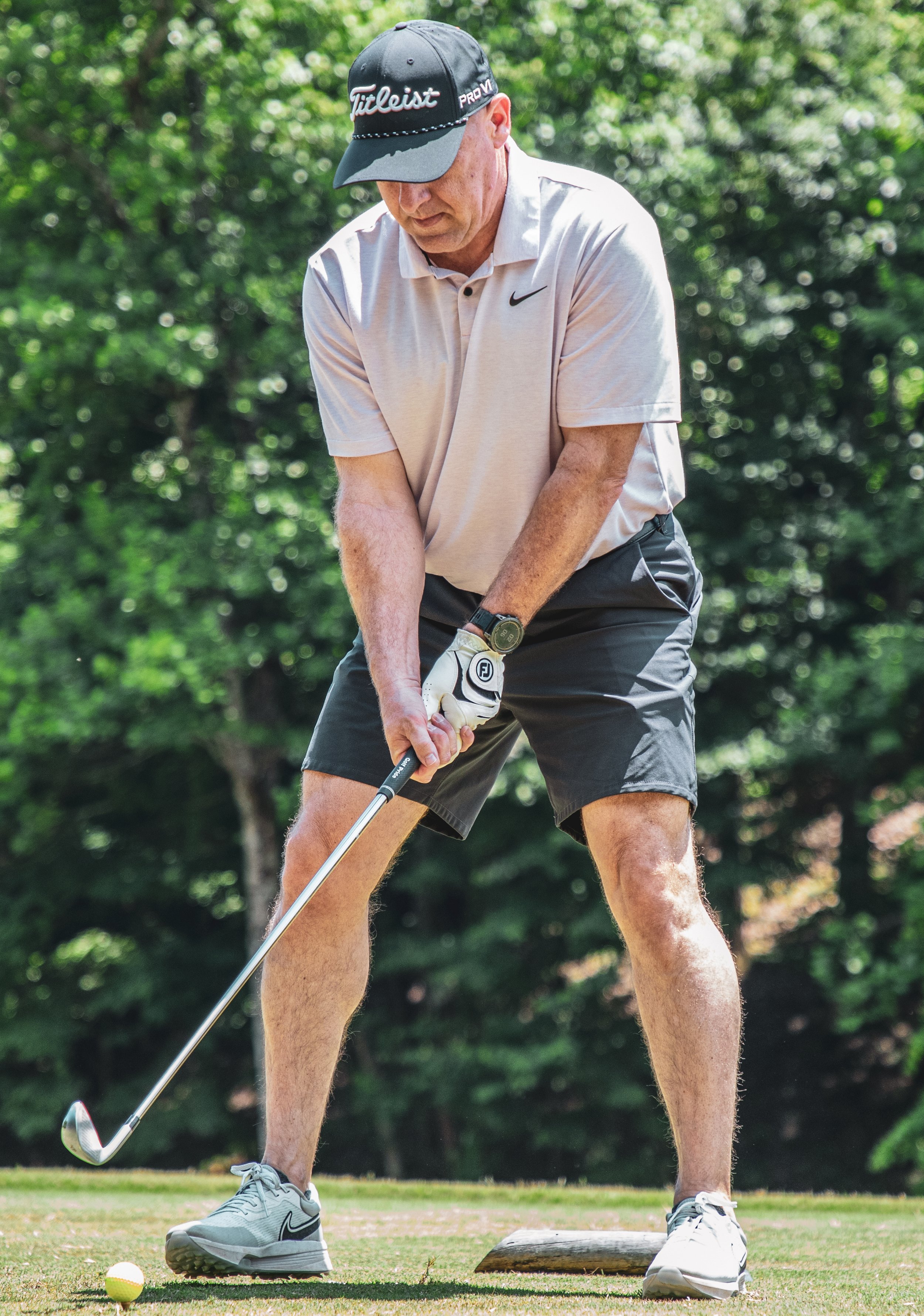 A man practicing golf, preparing to hit a golf ball on a tee, on a sunny day with a green, wooded background.