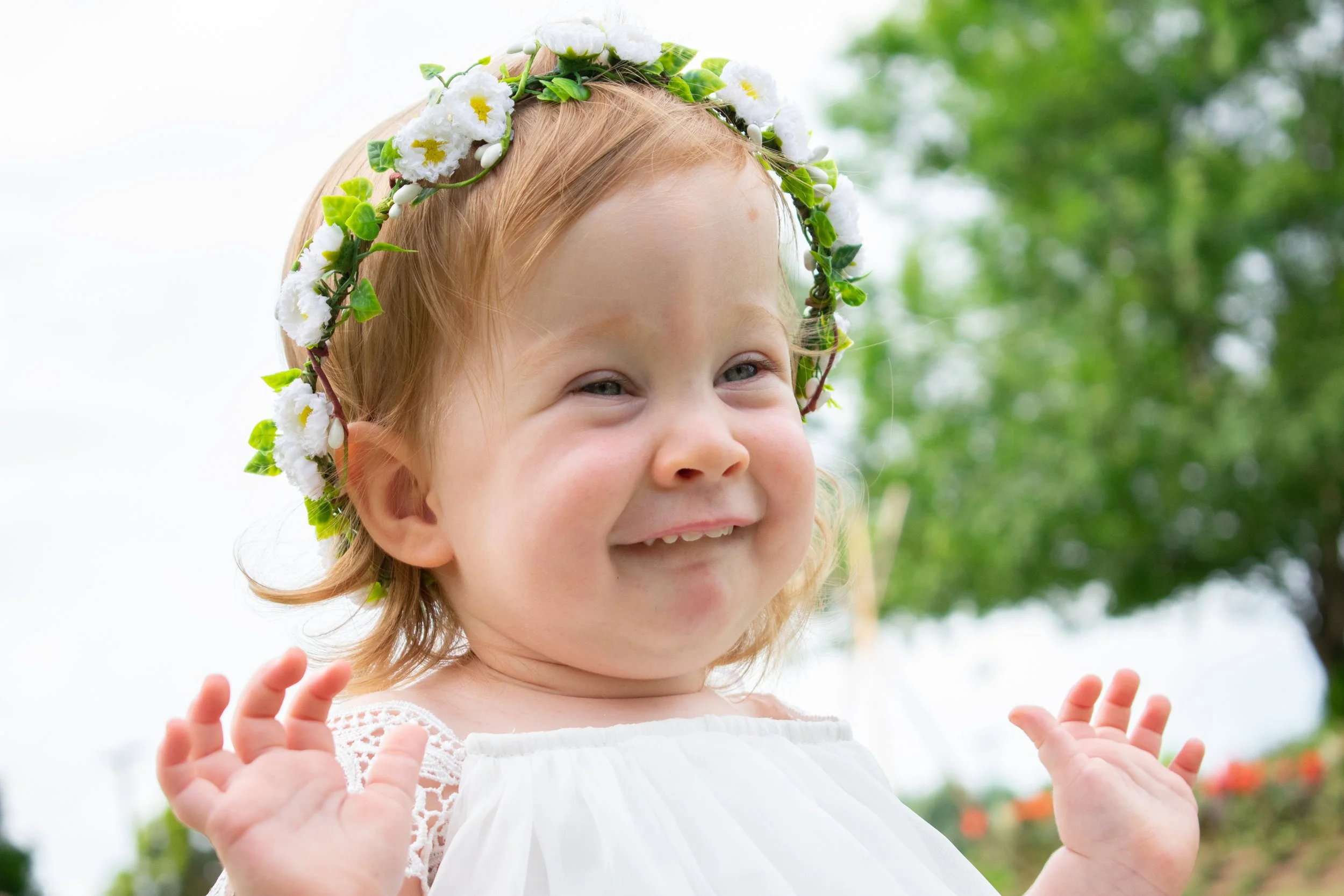 A smiling young girl with red hair wearing a white dress and a floral crown made of white flowers and green leaves, outdoors with a blurred green background.