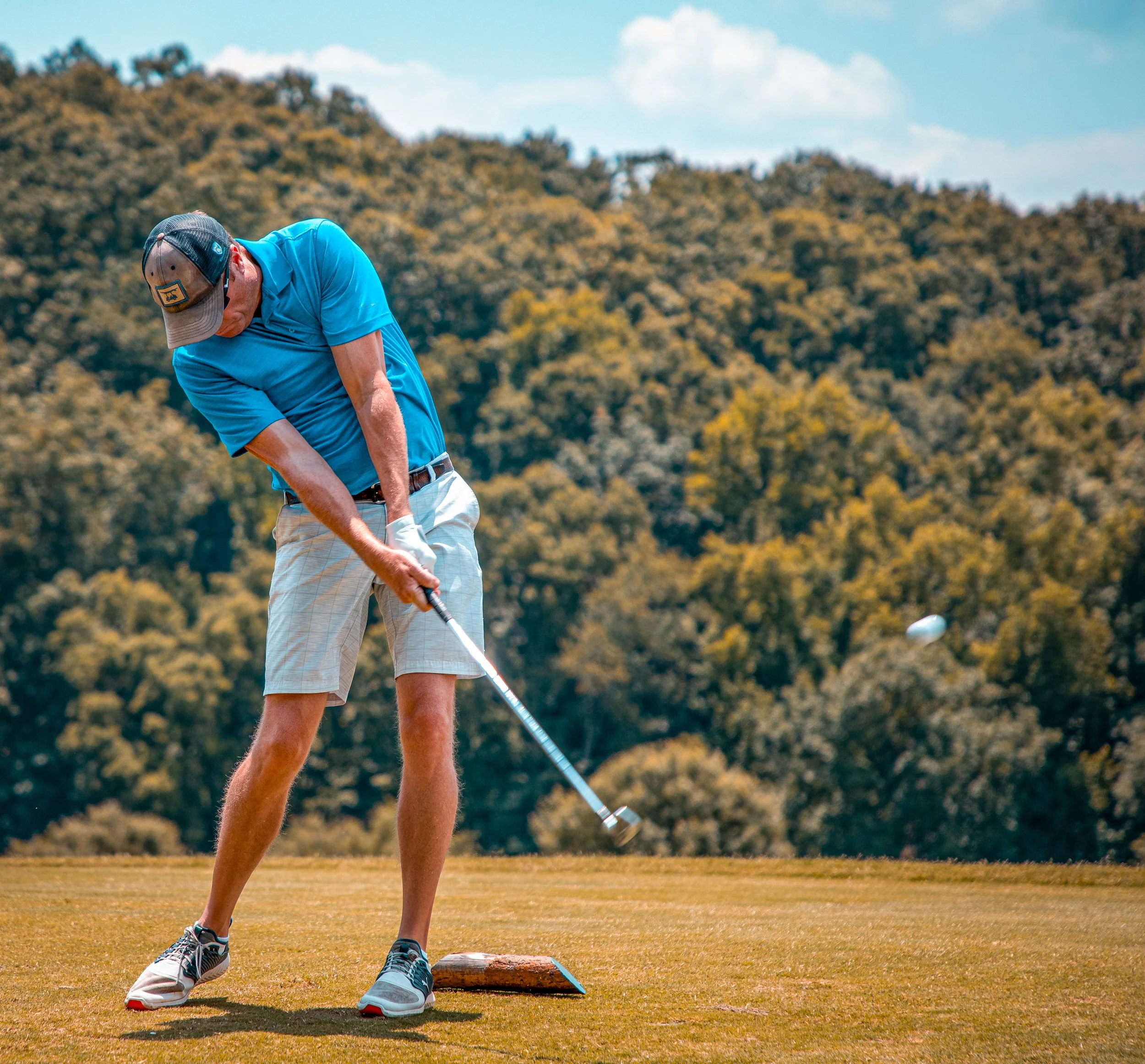A man in a blue polo shirt, beige shorts, and a baseball cap is setting up for a game of golf on a grassy course with trees and a cloudy sky in the background.