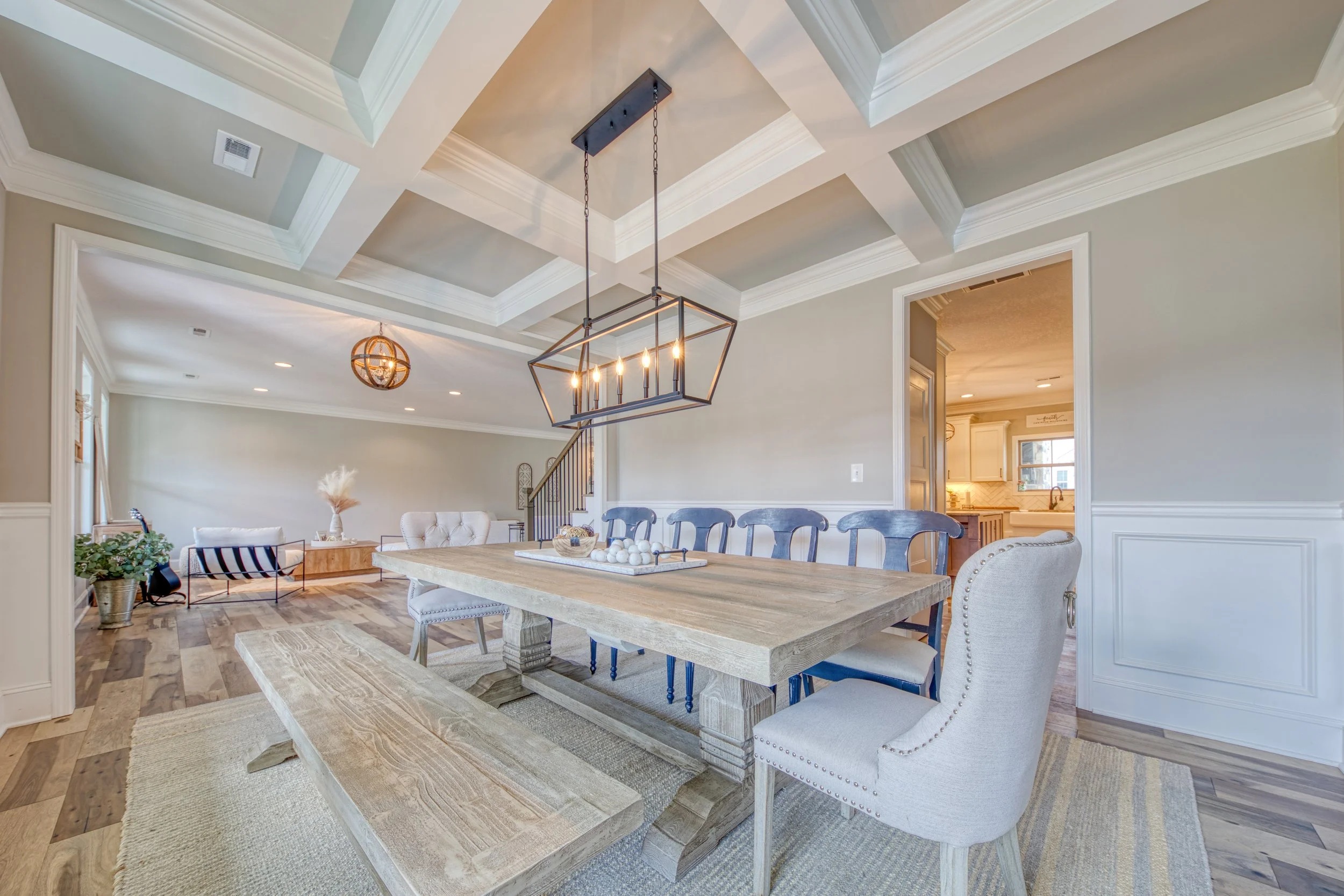 Dining room with a large wooden table, beige upholstered chairs, a modern geometric chandelier, and a coffered ceiling, with a view into a kitchen.