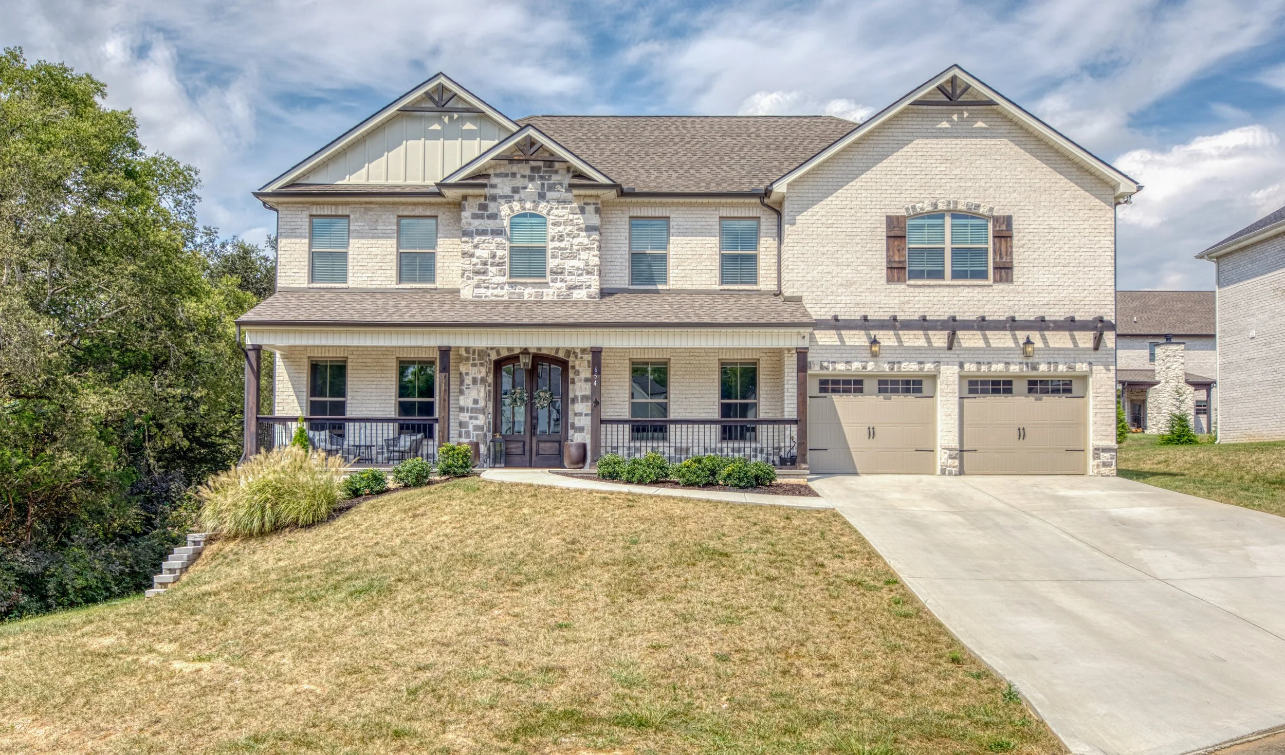 A two-story house with a brick and stone exterior, front porch, attached garage, and a well-kept lawn under a partly cloudy sky.