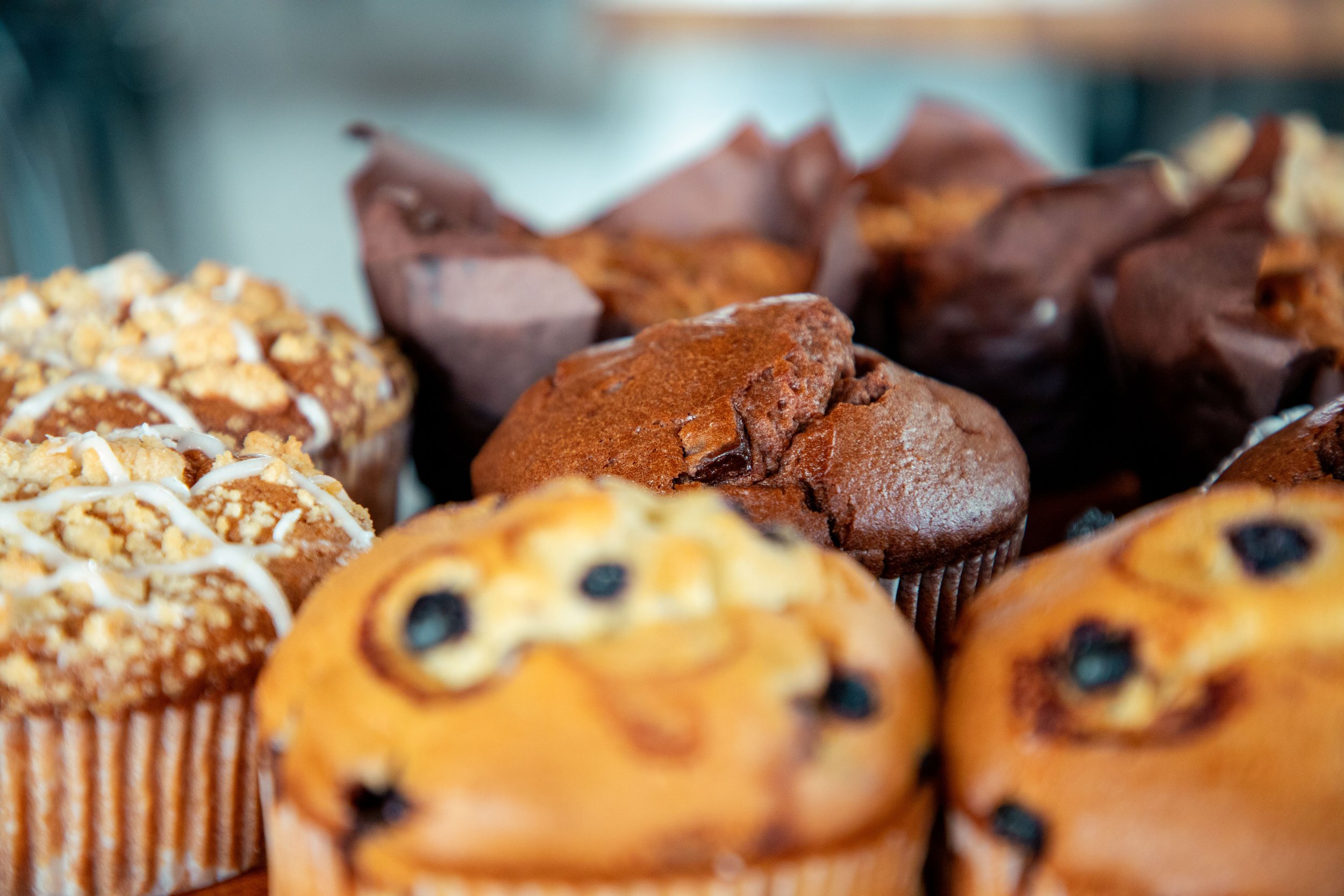 A close-up photo of a variety of baked muffins, including chocolate chip, crumb-topped, and possibly banana or other fruit flavors, displayed together.