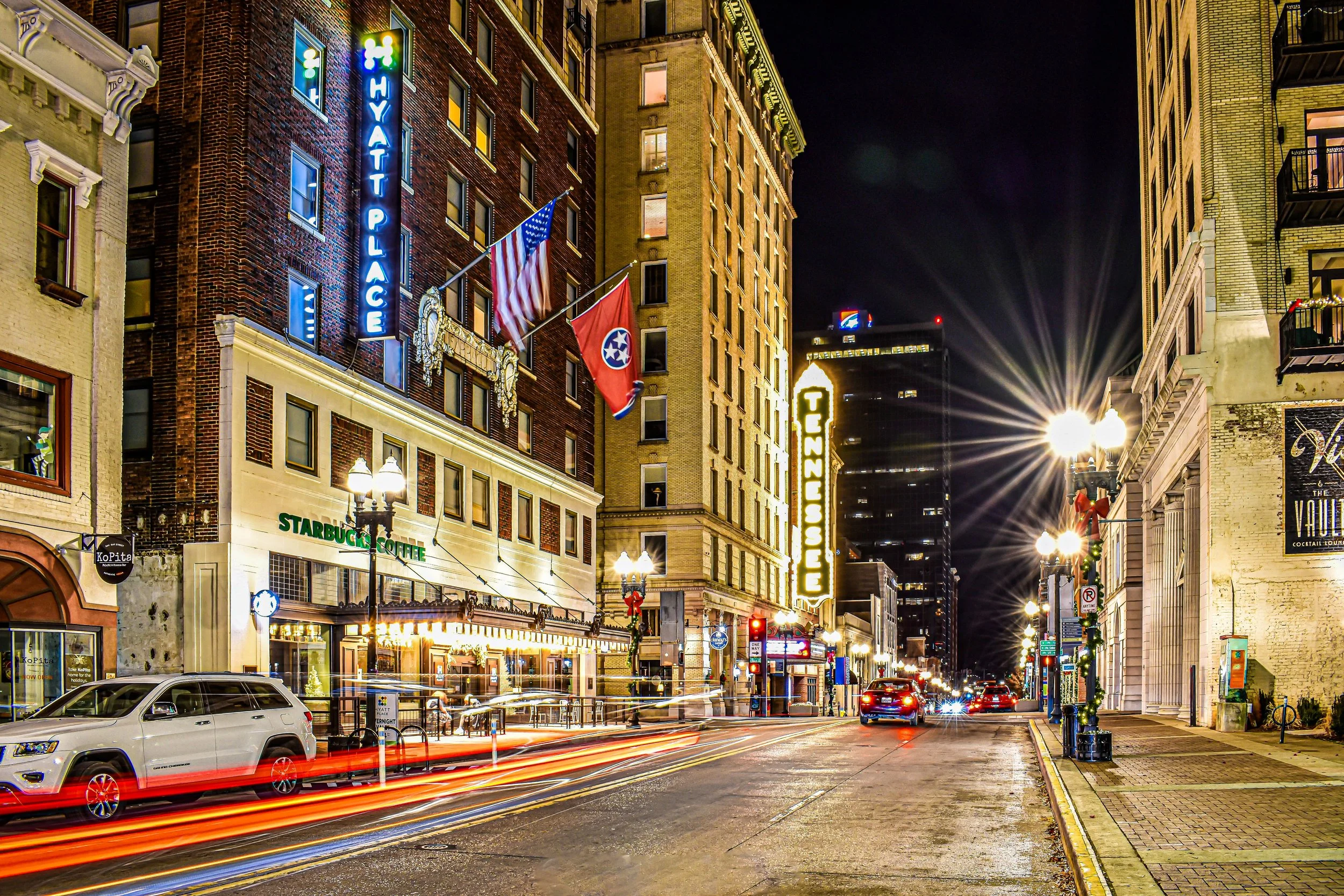 Nighttime street scene with lit buildings, neon signs, flags, and streaks of vehicle lights from long exposure in a downtown area.