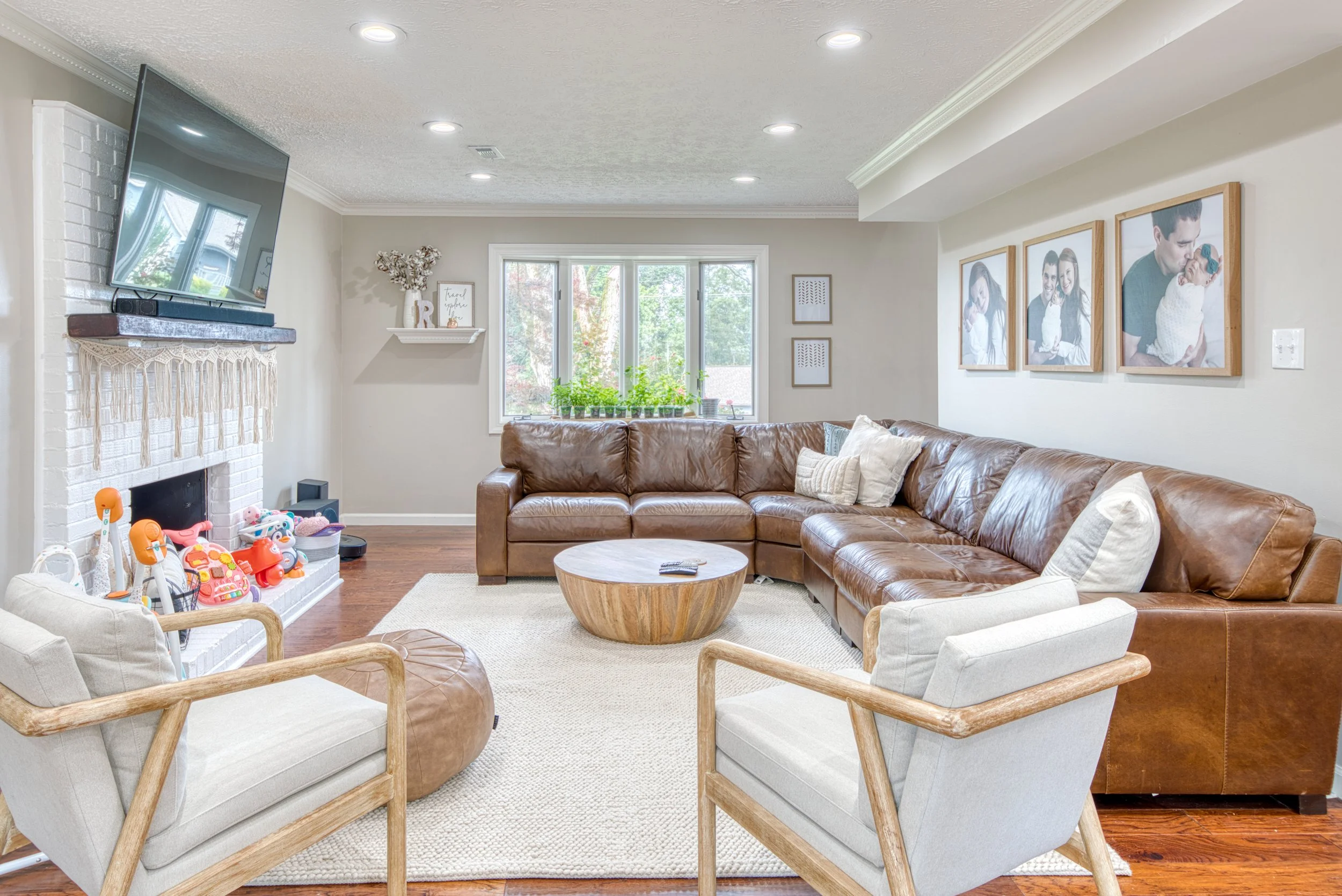 Bright living room with beige walls, a leather sectional sofa, and framed family photos on the wall.