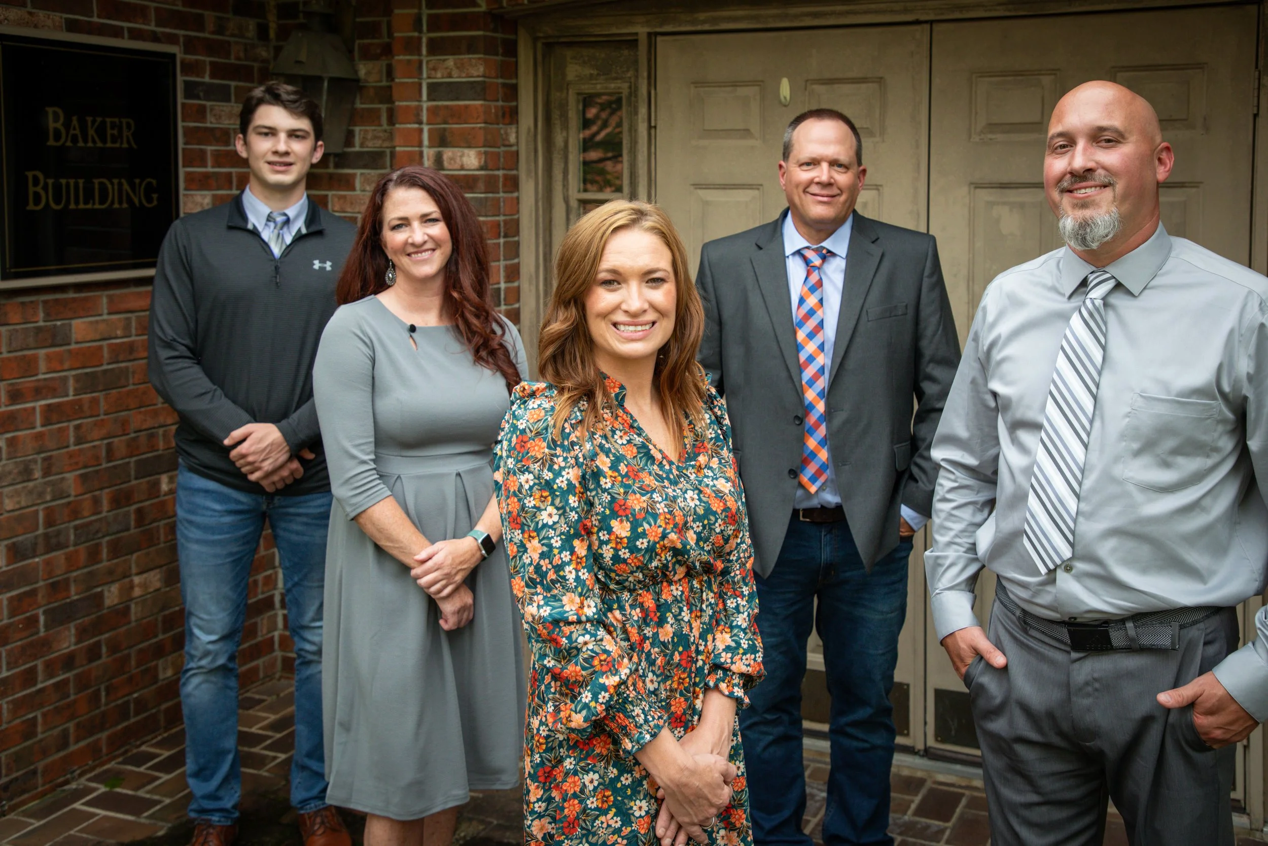 Group of five people standing outside near a brick building with a sign that says 'Baker Building'. They are smiling and dressed in business casual and formal attire.